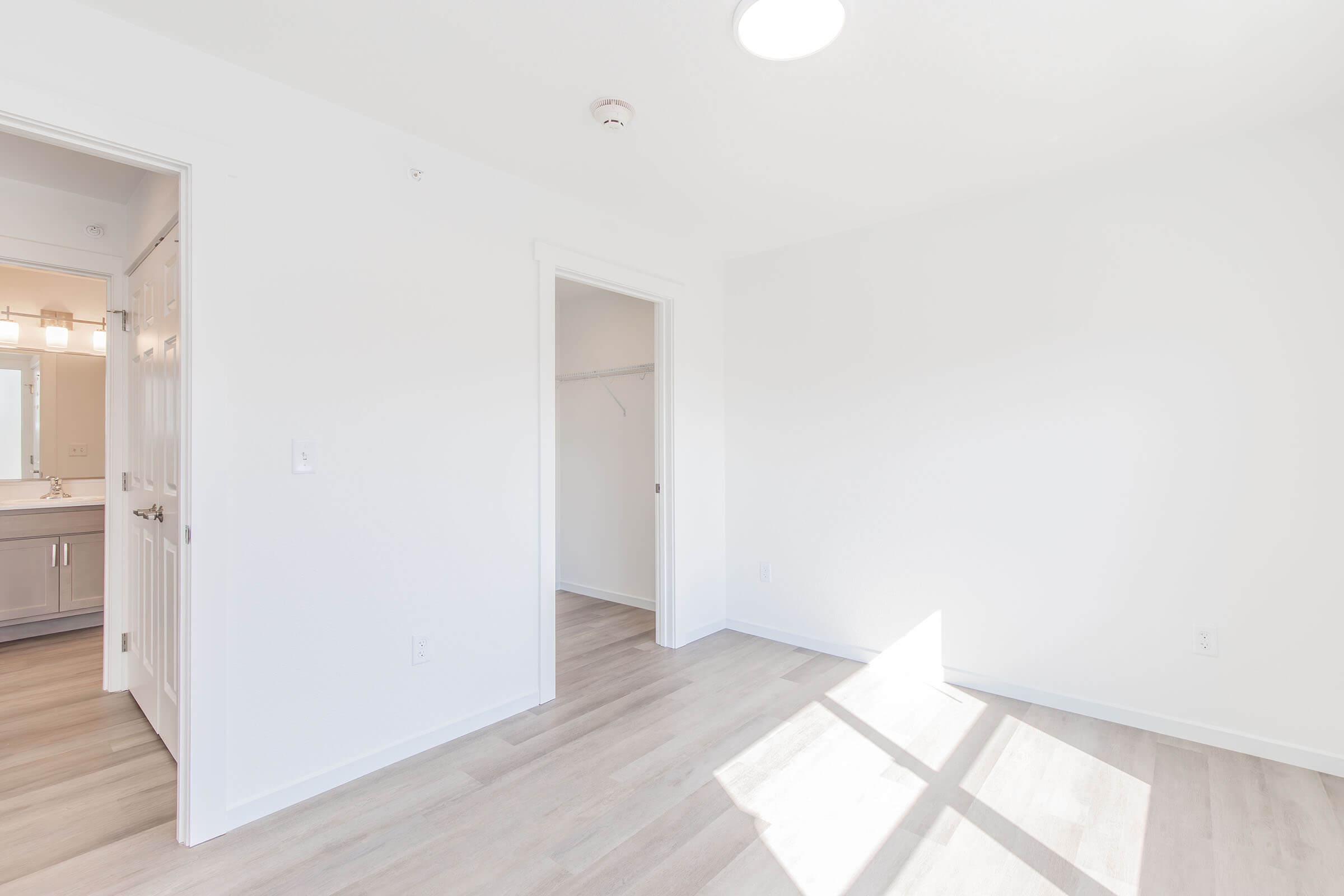 A bright, empty room with light-colored walls and wood laminate flooring. A sunlit patch on the floor indicates a window nearby. There is a door on the left leading to a bathroom and an open closet on the right. The overall ambiance is clean and modern.