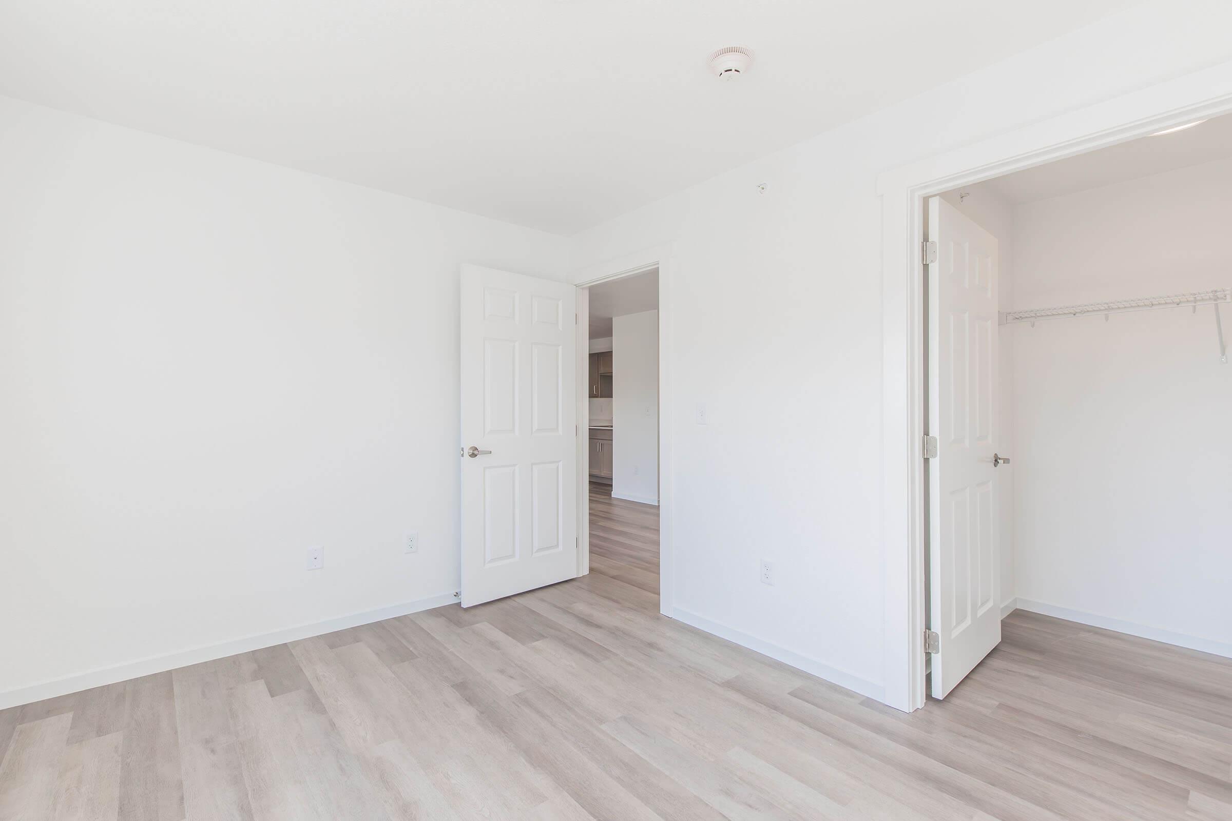 Empty room with light-colored walls and wooden flooring. Two open doors lead to adjacent spaces, while a closet with a hanging rod is visible to the right. Natural light brightens the space, creating a clean and minimalist look. The room is unfurnished, highlighting its potential for decoration.