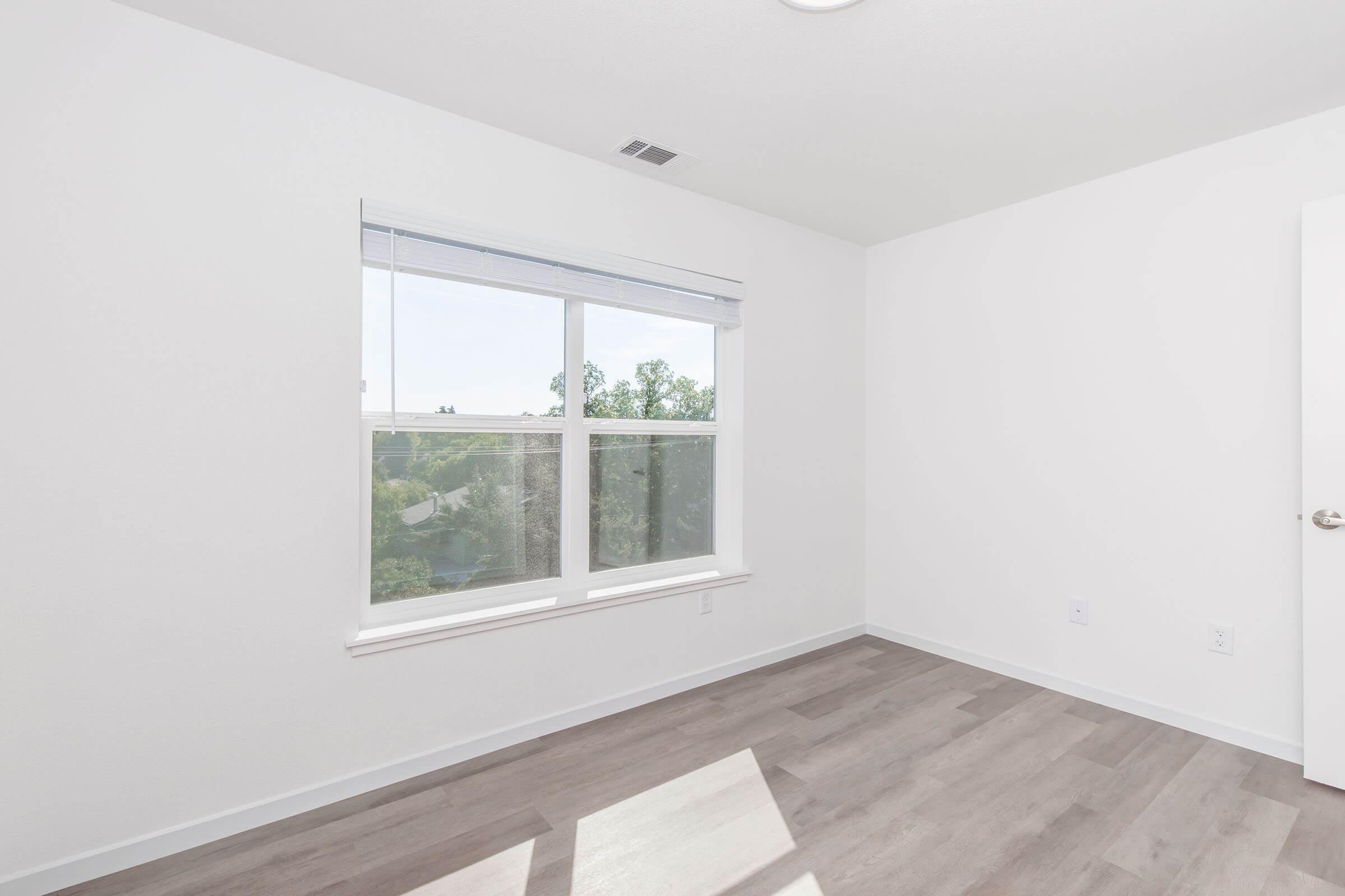 Bright, empty room with white walls and large window allowing natural light. The floor is light-colored wood or laminate, and there are no furnishings. The view outside the window includes greenery. The space appears clean and modern, suitable for various uses such as a bedroom or office.