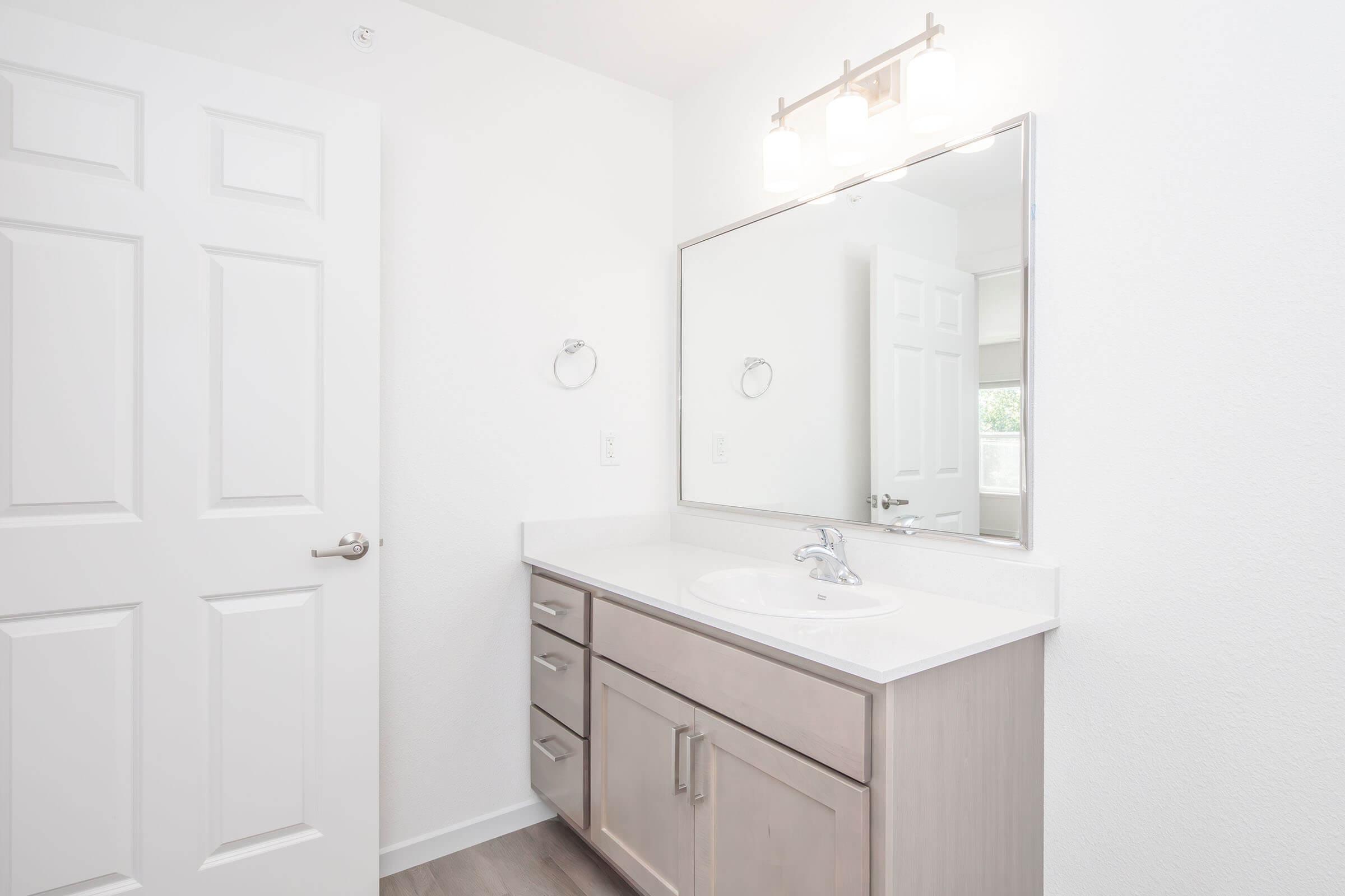 A modern bathroom featuring a light-colored vanity with drawers, a sleek white countertop, and a large mirror above the sink. The walls are painted white, and there is a door visible leading to another room. Soft lighting complements the bright, clean aesthetic.