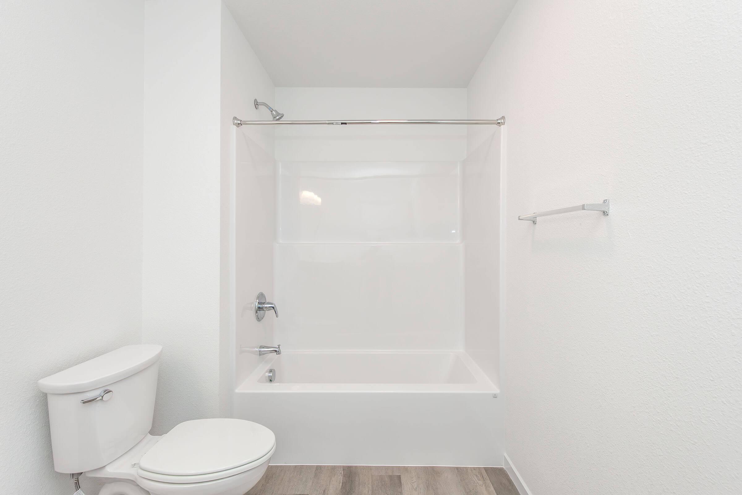 A clean, modern bathroom featuring a white bathtub with a glass shower curtain, a toilet, and a simple towel rack. The walls and fixtures are white, creating a bright and minimalist aesthetic, with light-colored flooring that complements the overall design.