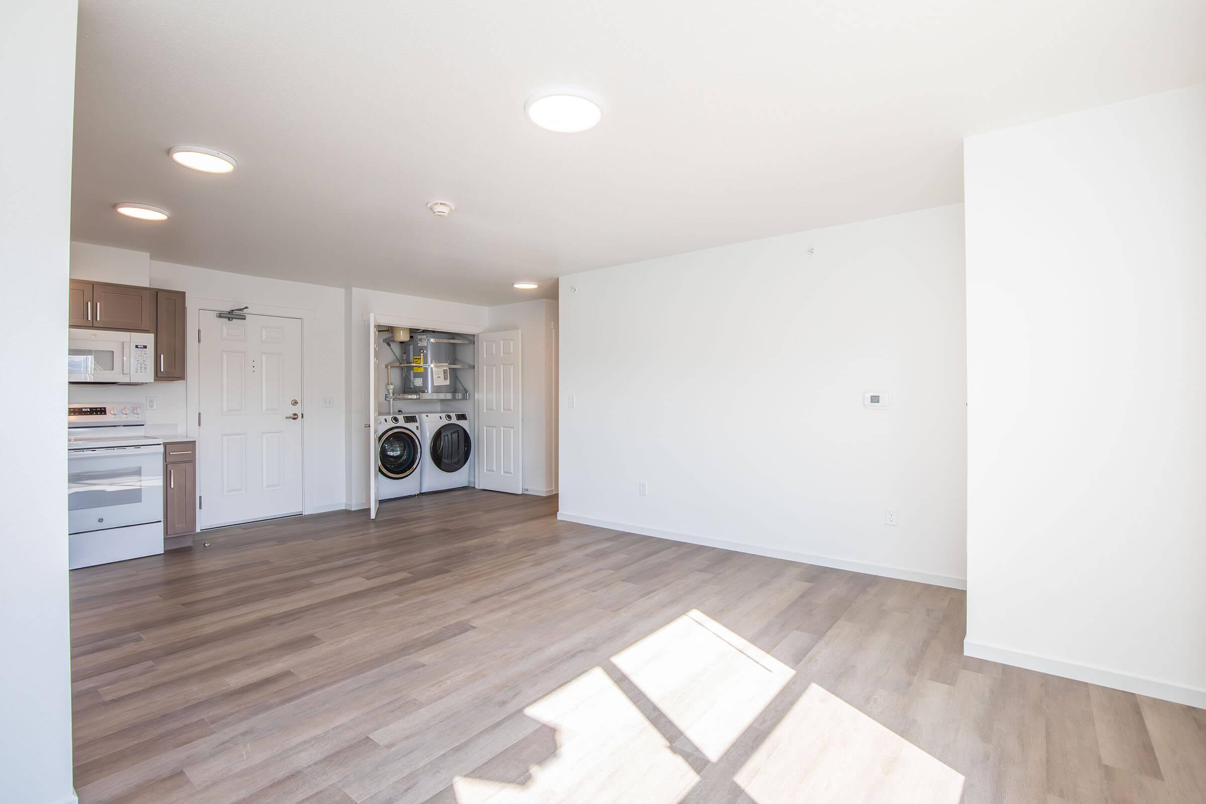 A spacious, modern living area with light-colored walls and hardwood floors. On the left, there is a kitchenette with appliances visible. Adjacent to it, a laundry area with a washer and dryer. Bright overhead lighting illuminates the room, creating an inviting and airy atmosphere.