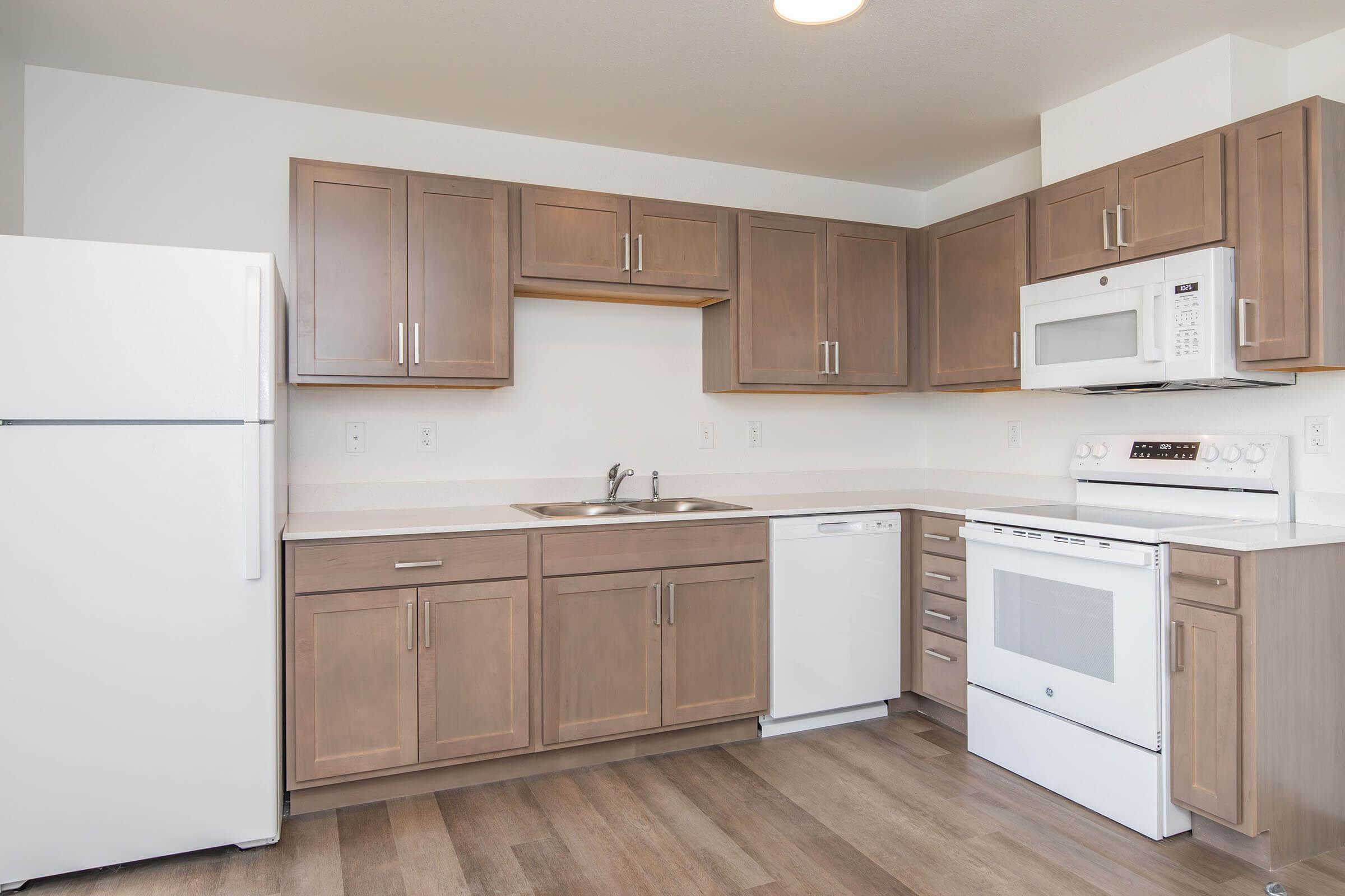 Modern kitchen featuring wooden cabinets, a stainless steel sink, and white appliances including a refrigerator, microwave, and dishwasher. The countertop is light-colored, and the flooring is a warm, neutral tone. The space is bright and well-organized.
