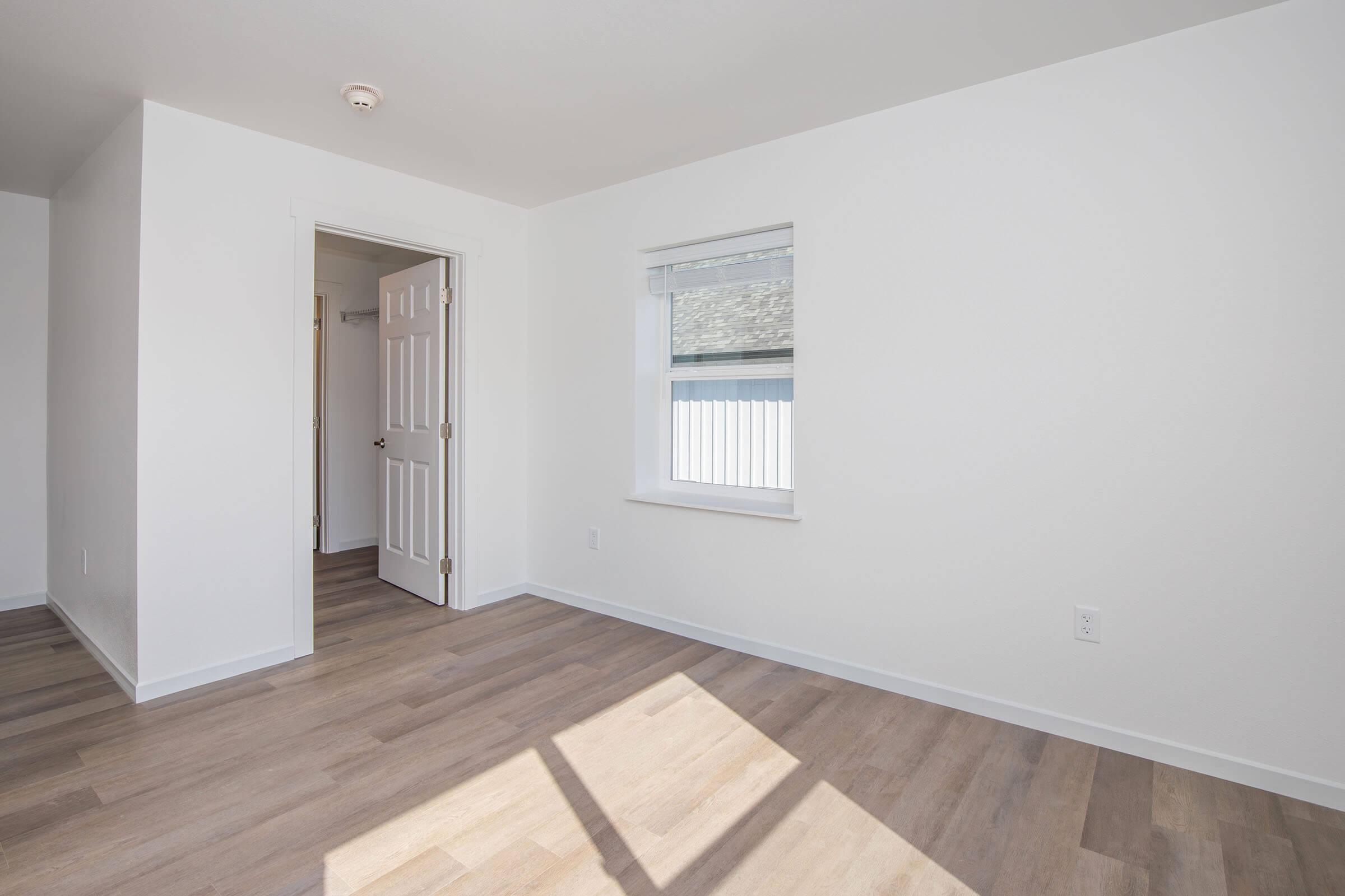 A bright, empty room with light-colored walls and light wood flooring. It features a closed door on the left, a window with a white blind, and light streaming in from the window. The space is clean and modern, emphasizing simplicity and openness.