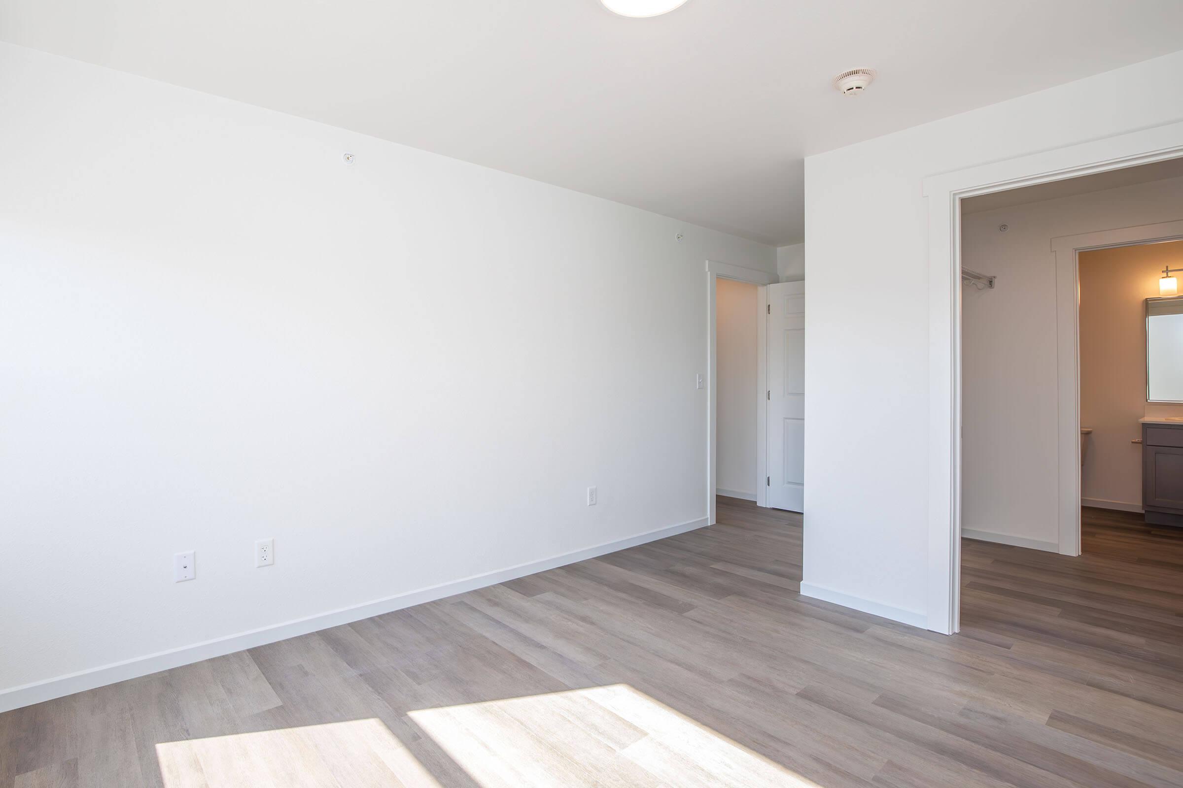 A bright, empty room with light-colored walls and wooden flooring. There is a doorway leading to another space, and a window allows natural light to enter. The room has minimal decor and a ceiling light fixture, creating an open and airy feel.