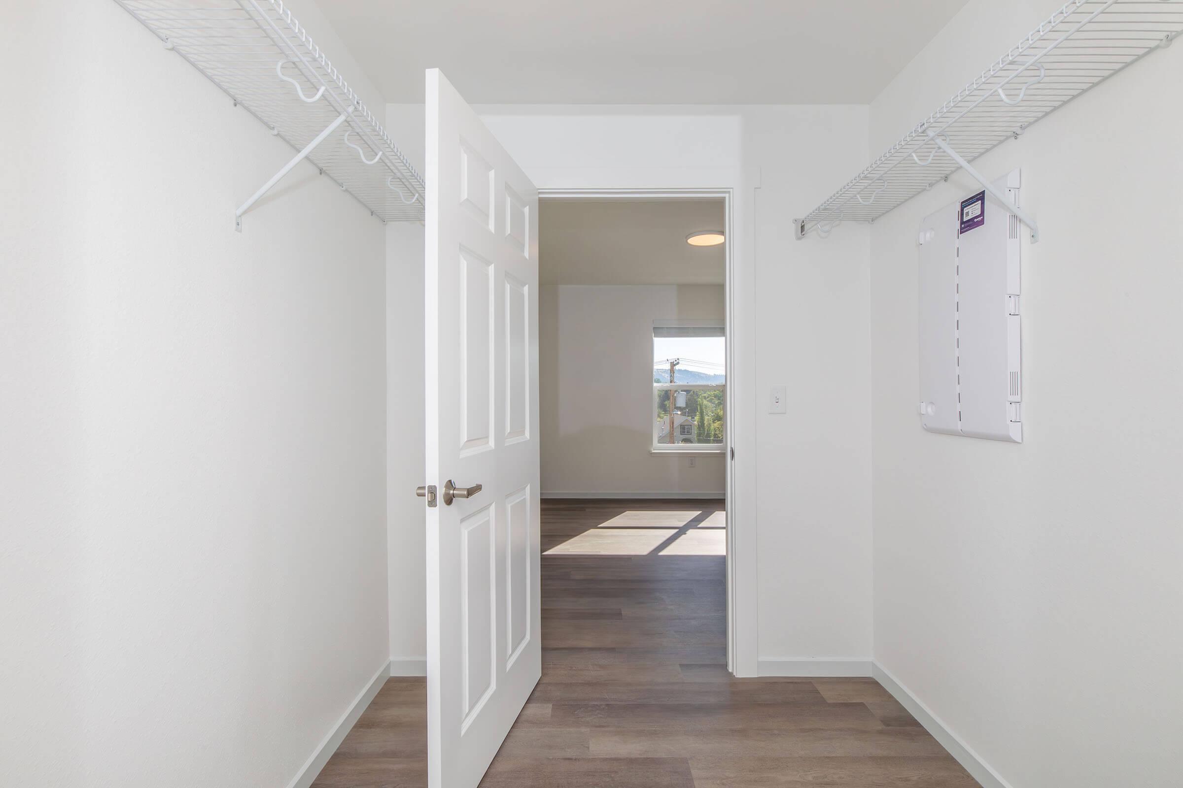 A spacious, well-lit closet with white walls and wooden flooring. The closet features wire shelving on both sides and an open door leading to a bright room beyond, with a window letting in natural light.