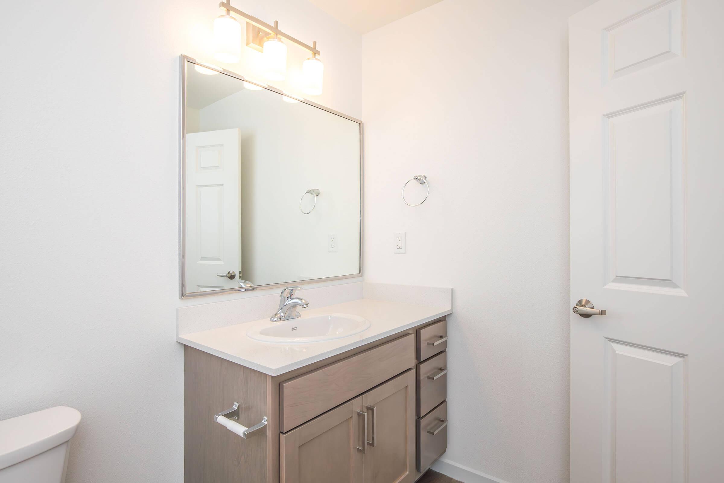 Bright and modern bathroom featuring a light-colored vanity with a sink, a large mirror above it, and illuminated by wall-mounted lights. A white door and towel ring are visible, along with a toilet in the corner. The walls are painted white, creating a clean and airy atmosphere.