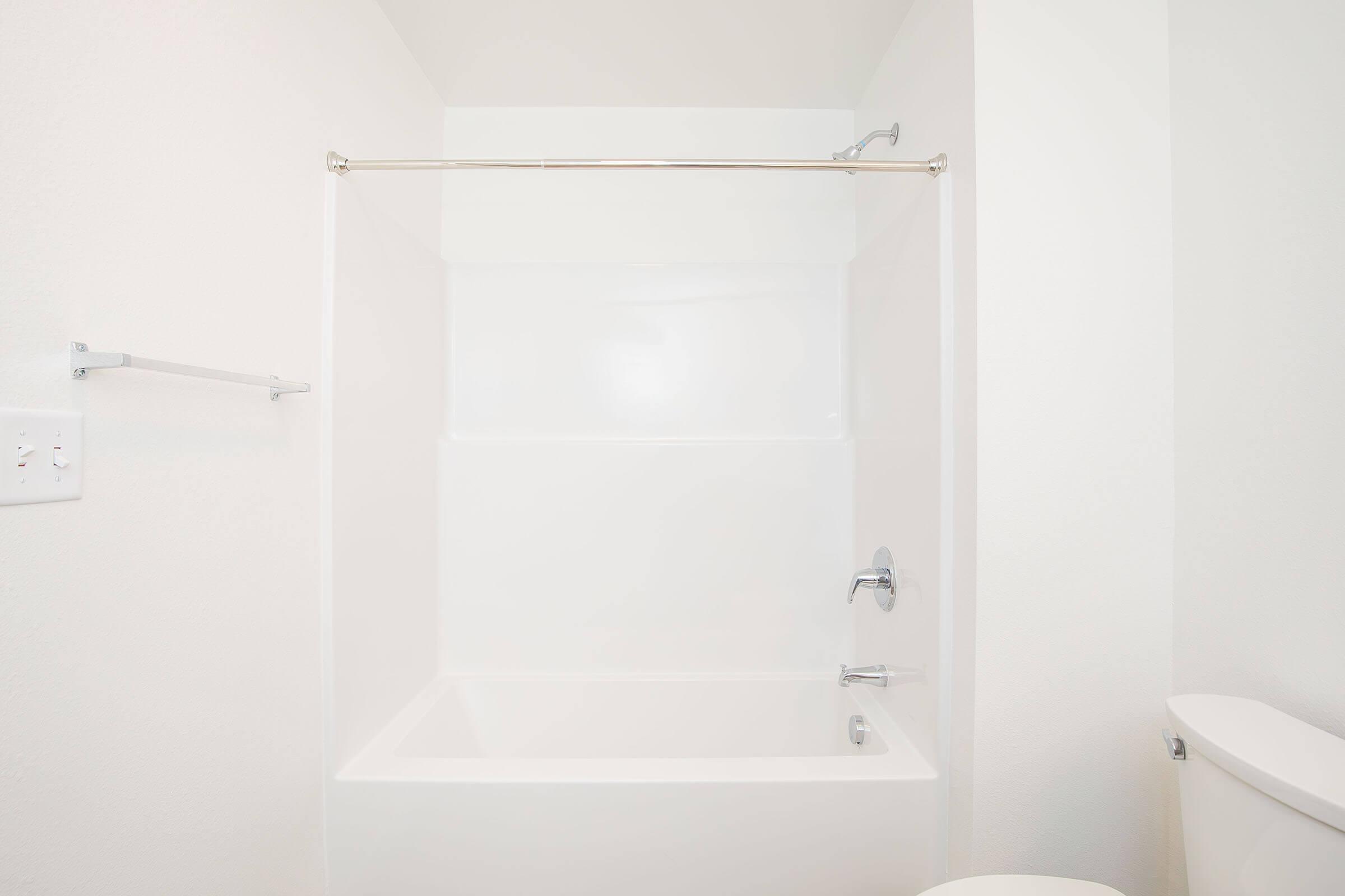 A clean, minimalist bathroom featuring a white bathtub with a shower rod and transparent curtain. The walls are also white, creating a bright and spacious appearance. A faucet is visible on the wall, and a toilet can be seen in the corner of the image.