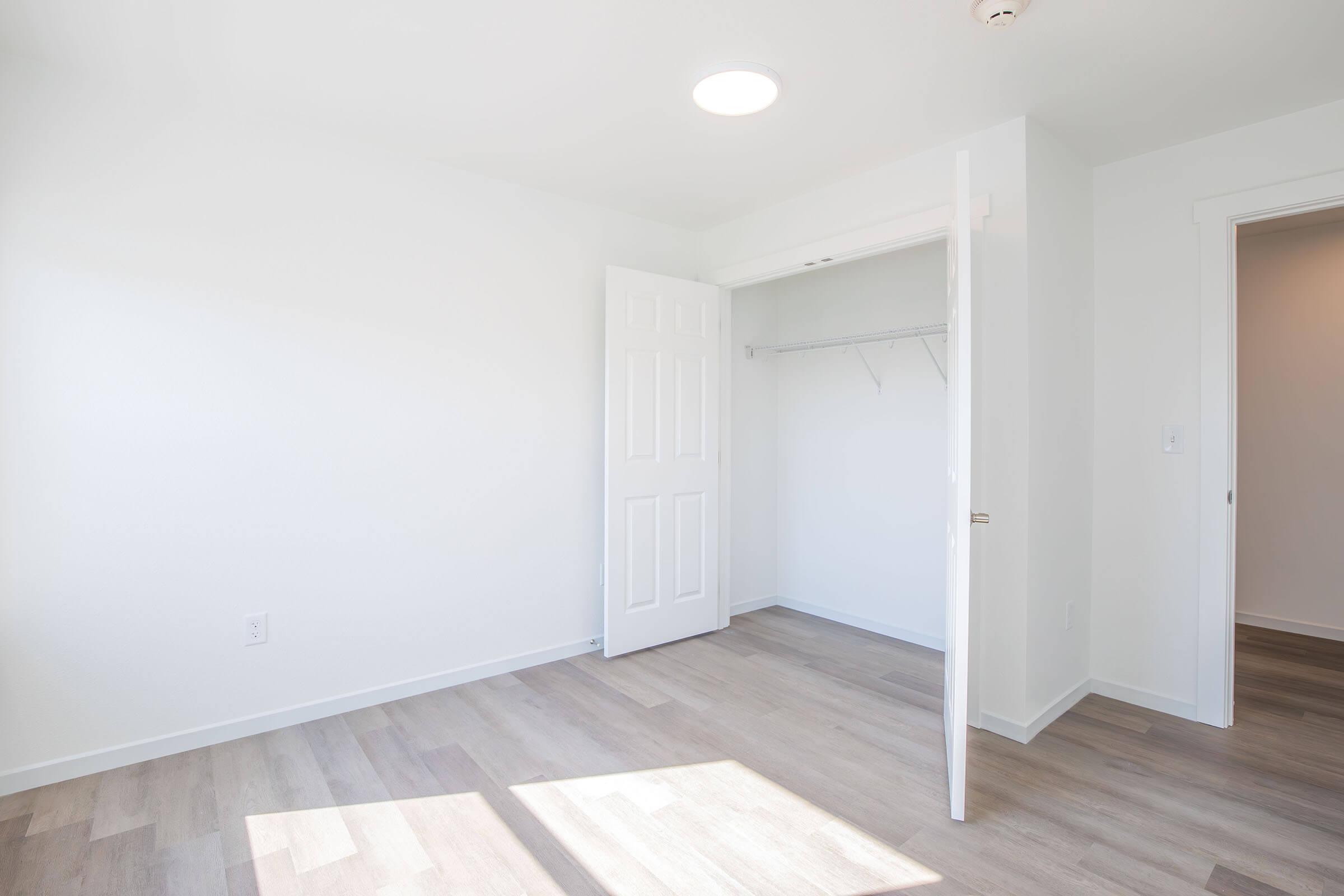 A bright, empty room with white walls and light-colored flooring. A closet with an open door is visible on the left, while sunlight filters through a circular ceiling light. There are no furnishings, creating a spacious and airy feel.