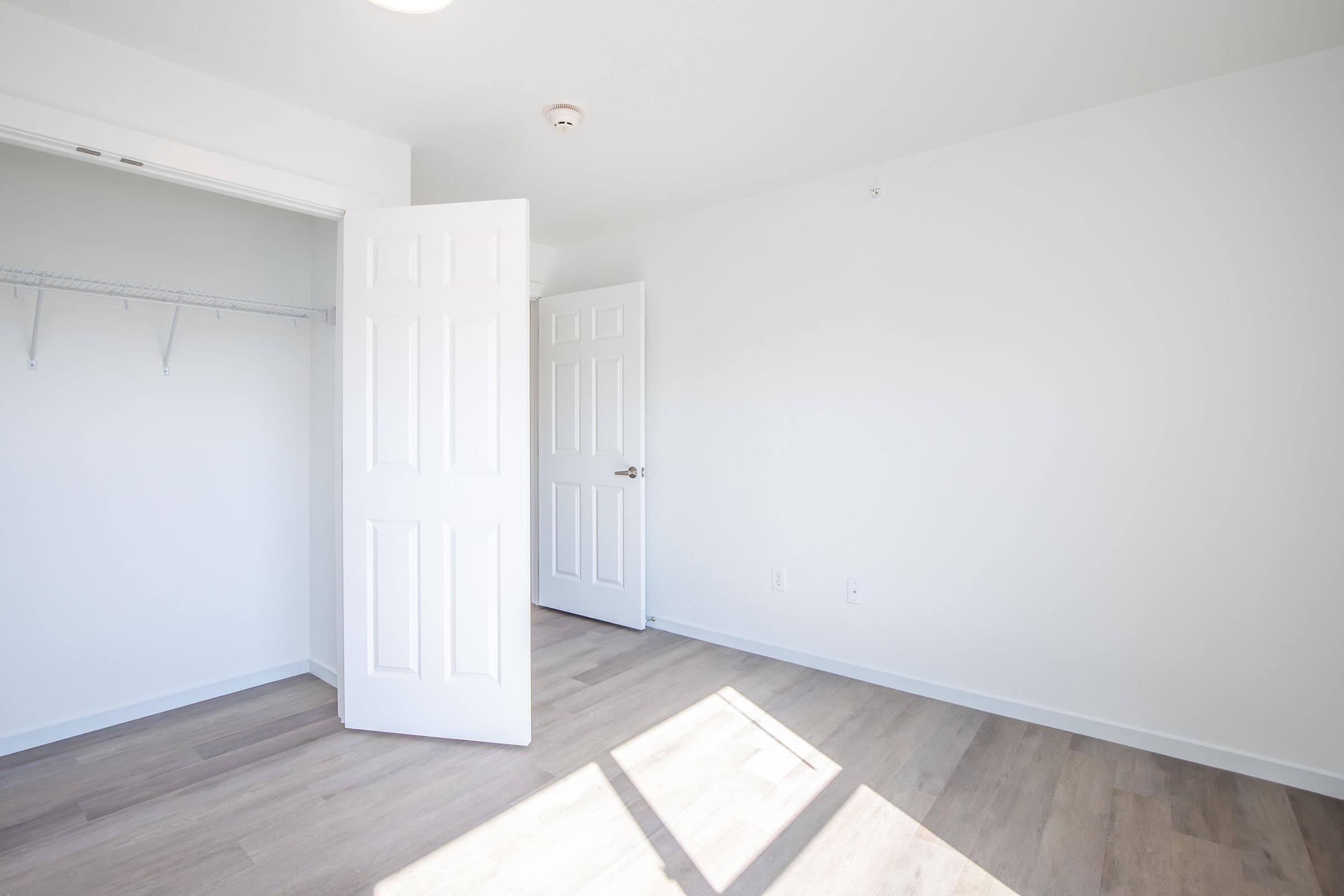 A bright, empty room with light-colored walls and wooden flooring. One door is open, leading to a closet with shelves, while another closed door is visible on the opposite wall. Sunlight streams in, creating a clean and spacious atmosphere.