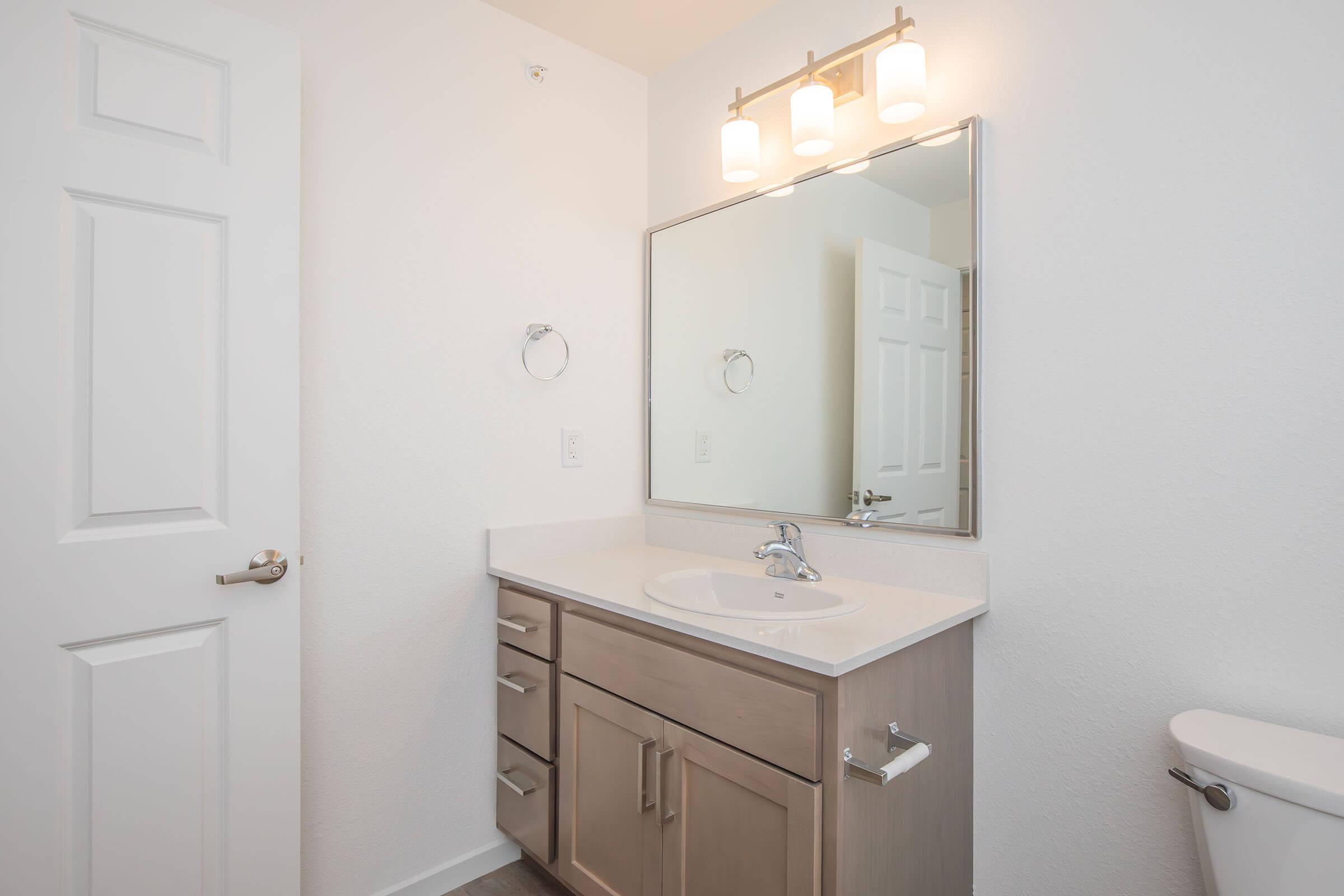 A modern bathroom featuring a single vanity with a sink, a large mirror above, and a light fixture with three bulbs. The cabinetry is light-colored wood, and there's a toilet positioned to the right. The walls are painted white, and the overall design is clean and minimalistic.