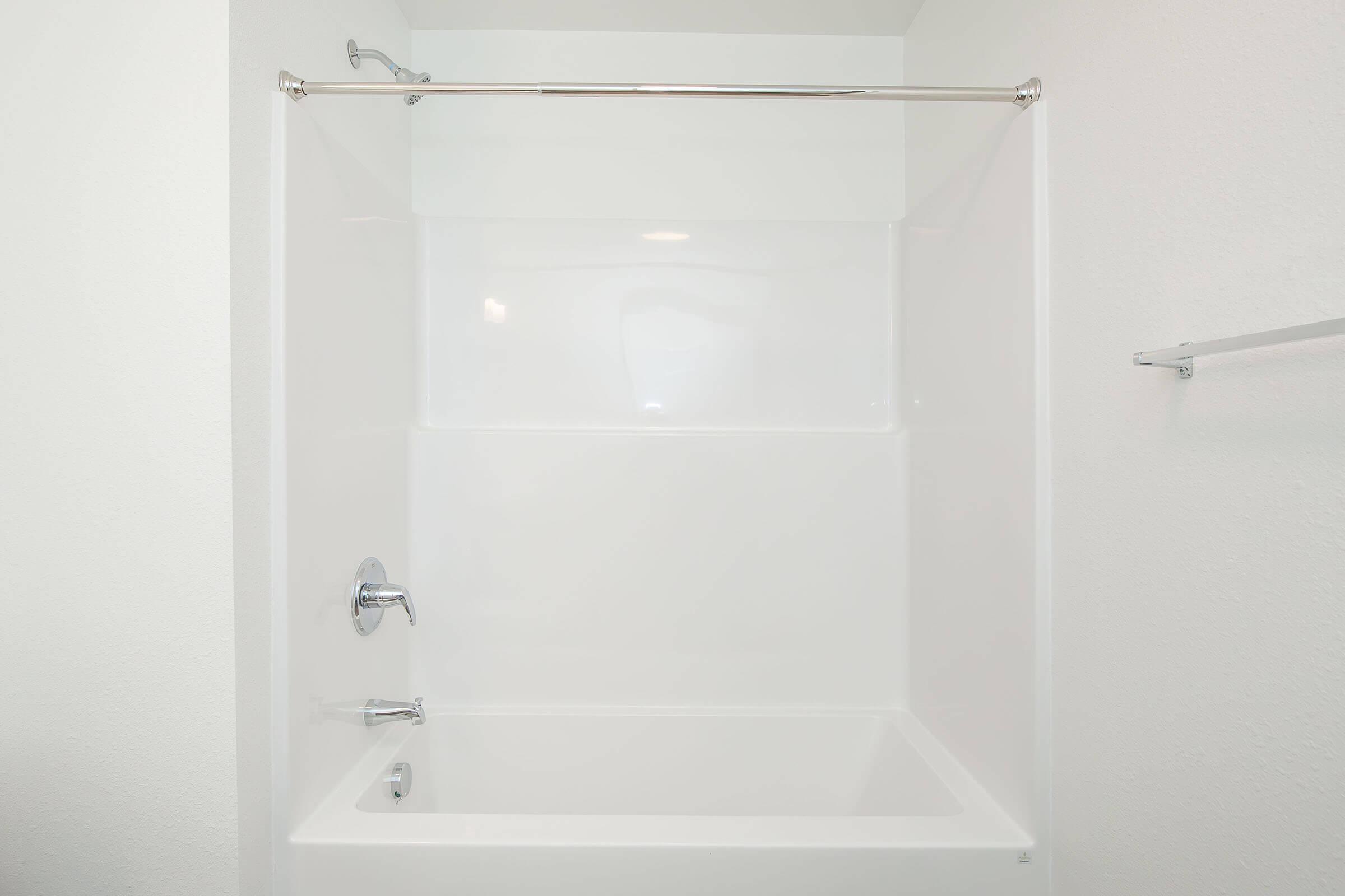A clean, modern bathtub and shower area with a white, smooth surface. It features a chrome faucet, a handheld showerhead, and a simple, minimalist design. The walls are unadorned and bright, emphasizing a fresh and uncluttered look.