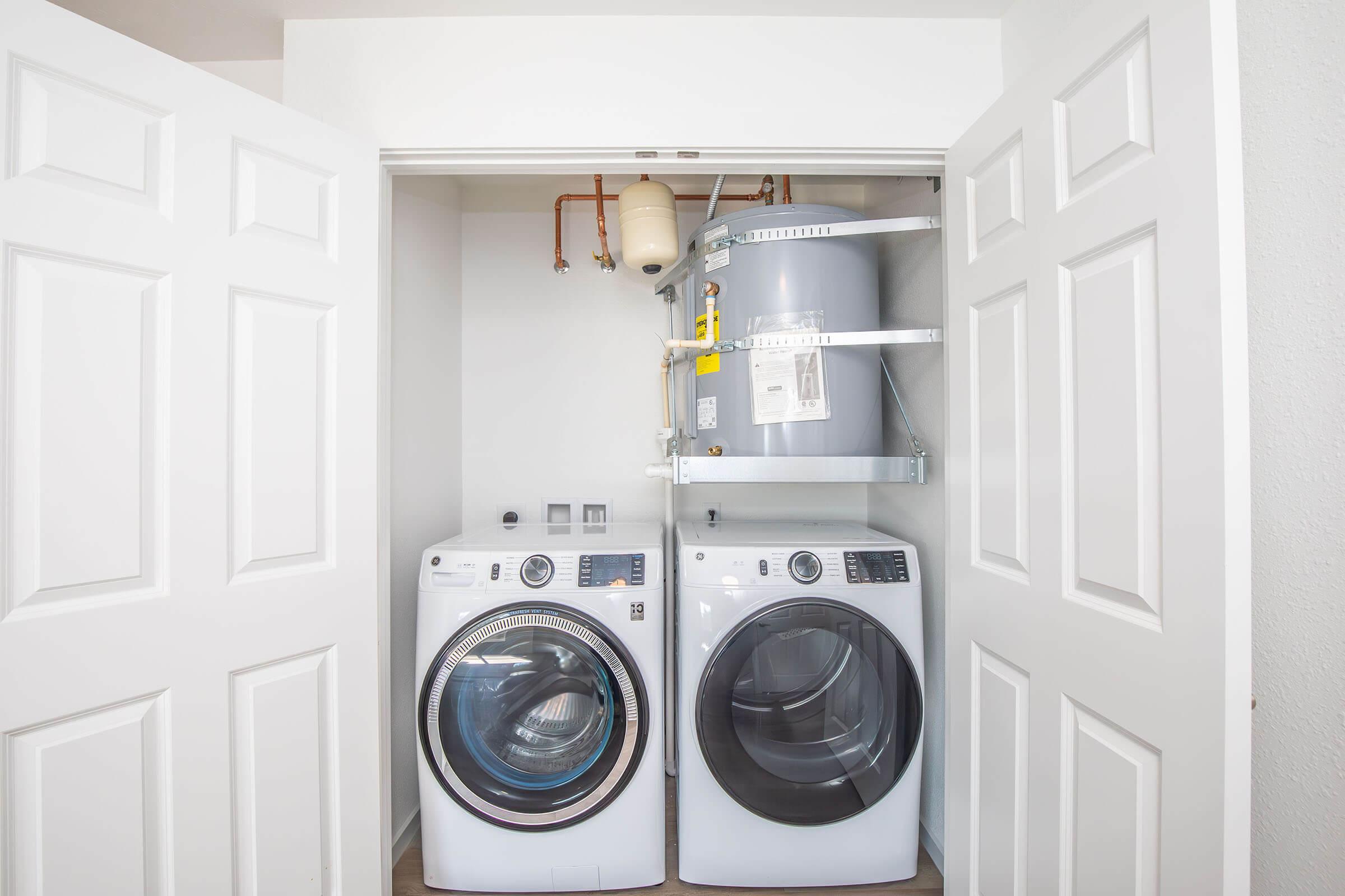 A compact laundry area featuring a stacked washer and dryer set in front of a water heater. The space is enclosed by double doors, and the appliances are modern and white, providing a clean and organized look. A shelf is installed above the water heater for additional storage.