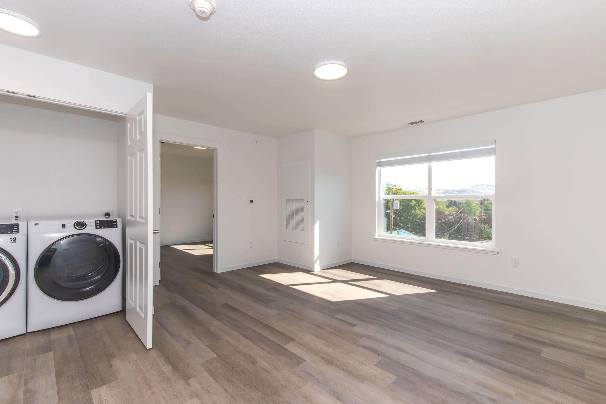 A bright, modern laundry room featuring white washing machines and dryers on the left. Sunlight floods in through a large window with a view of distant hills, illuminating the space with light hardwood flooring, neutral walls, and an open layout leading to another room.
