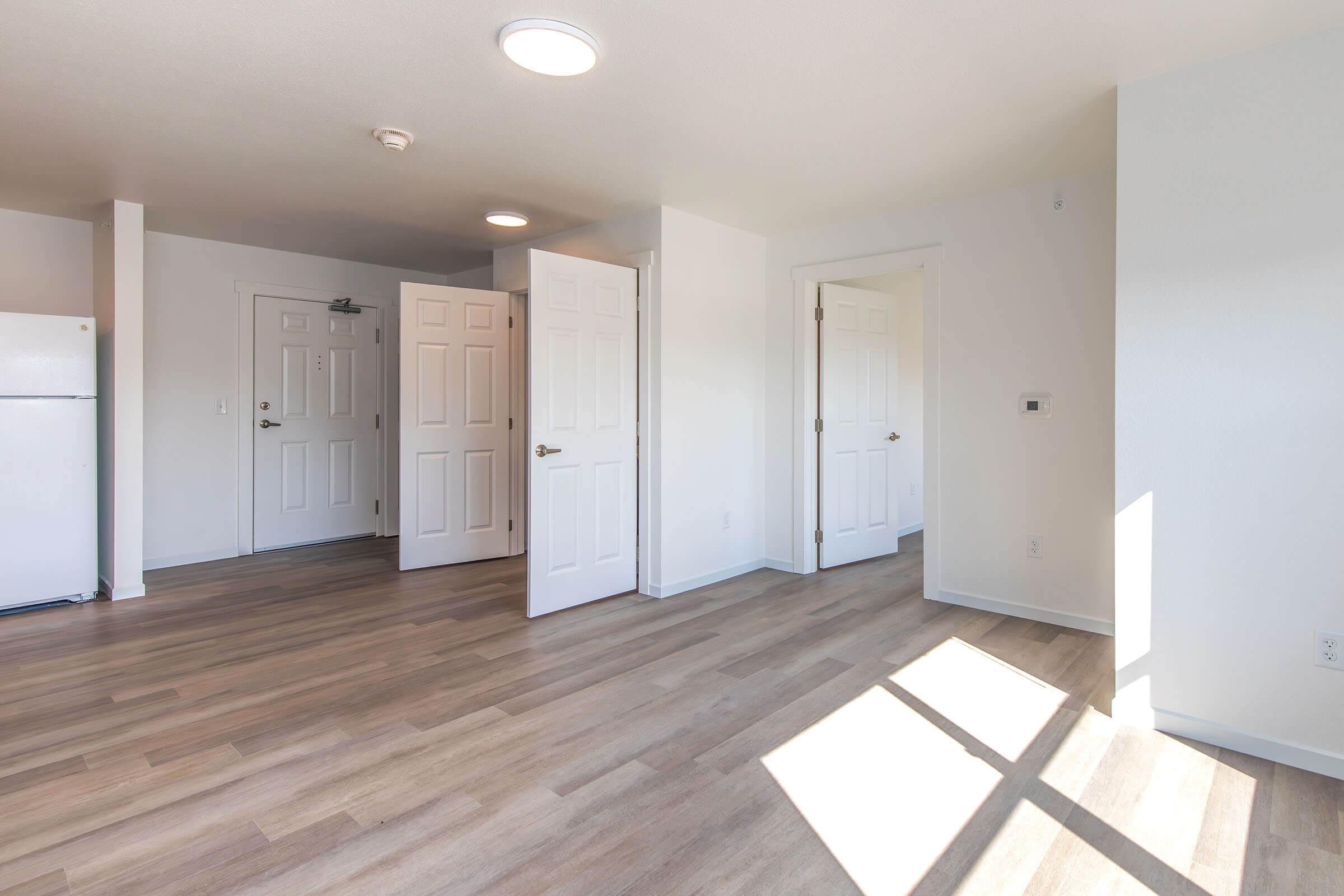 A bright, empty room with light-colored walls and wood-style flooring. A white refrigerator is visible in the corner. There are multiple doorways leading to other areas, one of which has an open door. Natural light flows in from a window, creating a spacious feel.
