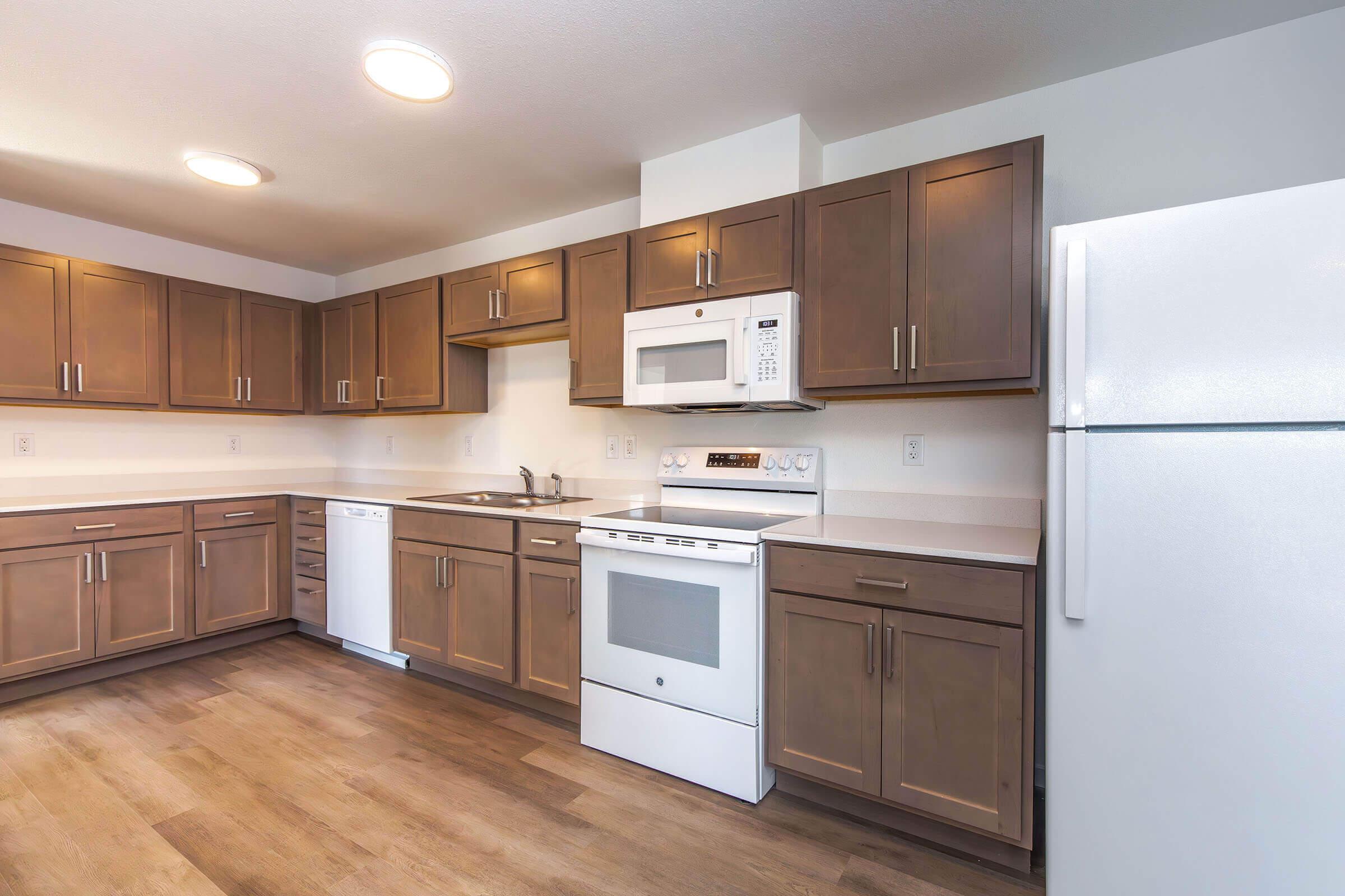 Modern kitchen featuring wooden cabinets, white countertops, and stainless steel appliances. Includes a refrigerator, microwave, and stove, with a sink and dishwasher. The floor has a warm wood finish and the space is well-lit with overhead lighting.