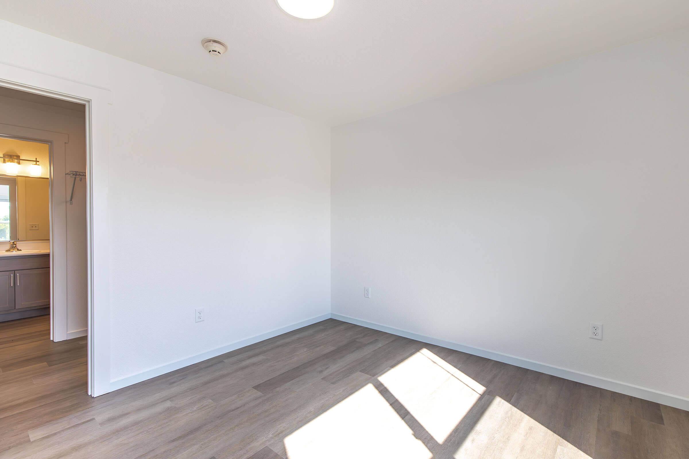 Empty room with light-colored walls and wooden flooring. A door on the left leads to a bathroom area, while sunlight streams in through a window, creating a bright and open atmosphere. The room lacks furniture, emphasizing its spaciousness.