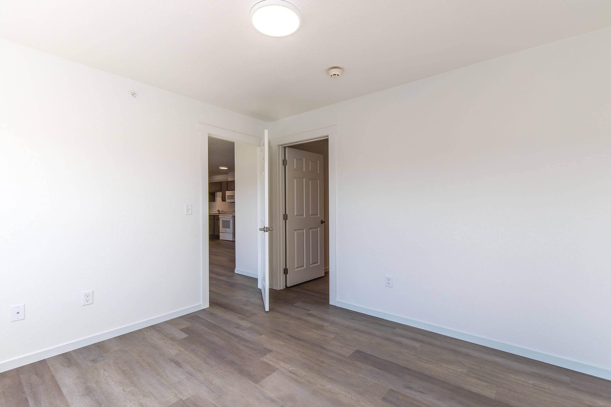 A well-lit, empty room with light-colored walls and wood laminate flooring. A partially opened door leads to another room or area. The space appears clean and modern, showcasing a simple design with ample natural light.