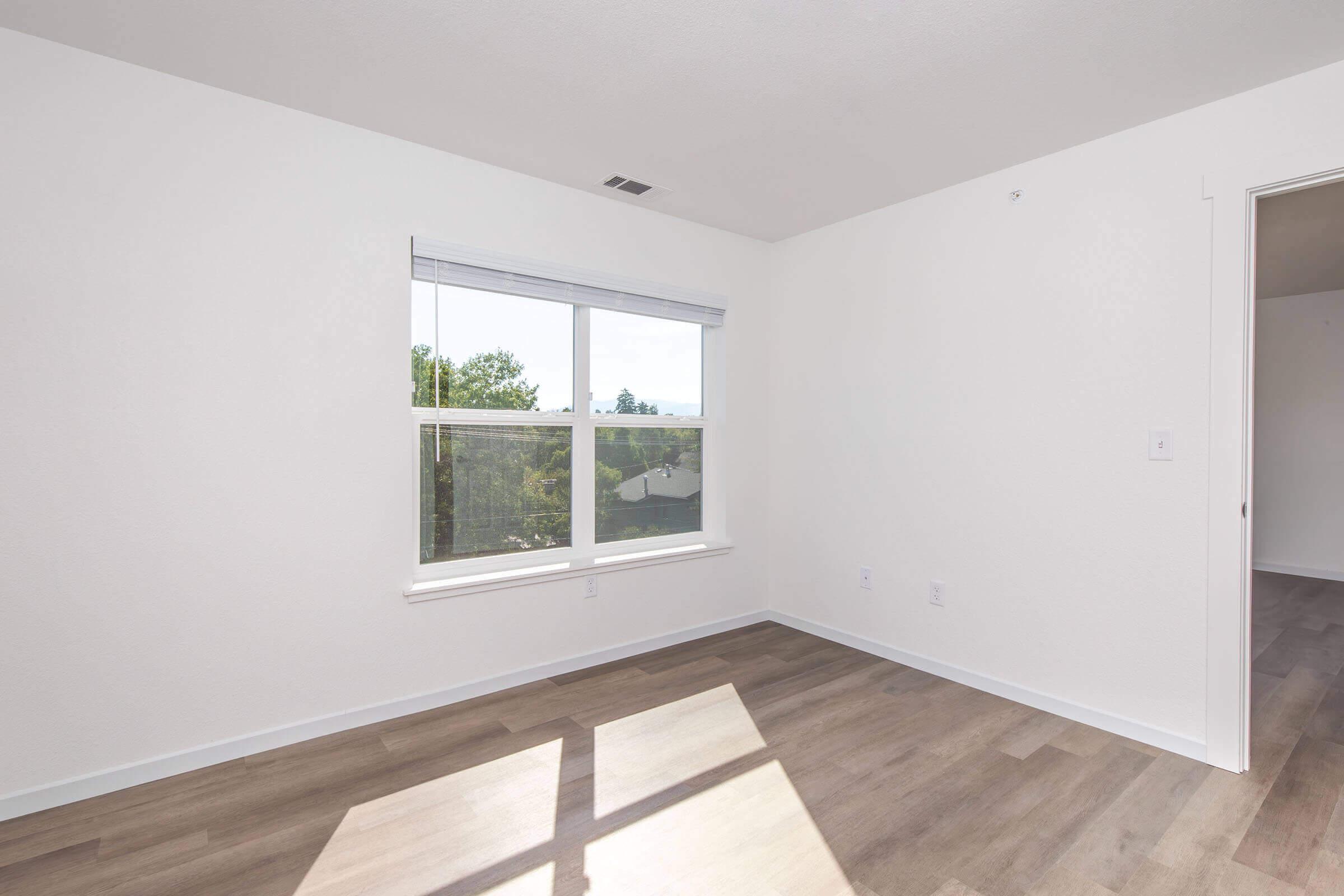 A bright, empty room with light-colored walls and a large window letting in natural light. The floor is light hardwood, and there are no furnishings. A door is visible in the corner, suggesting entry to another space.