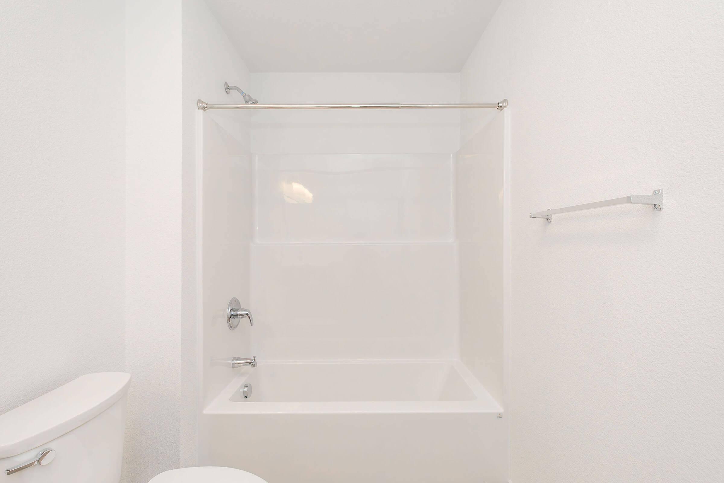 A clean, empty bathroom featuring a white bathtub with a shower curtain rod, a modern faucet, and a white toilet. The walls and ceiling are smooth and painted white, creating a bright, spacious ambiance. A towel rack is mounted on the wall opposite the bathtub.