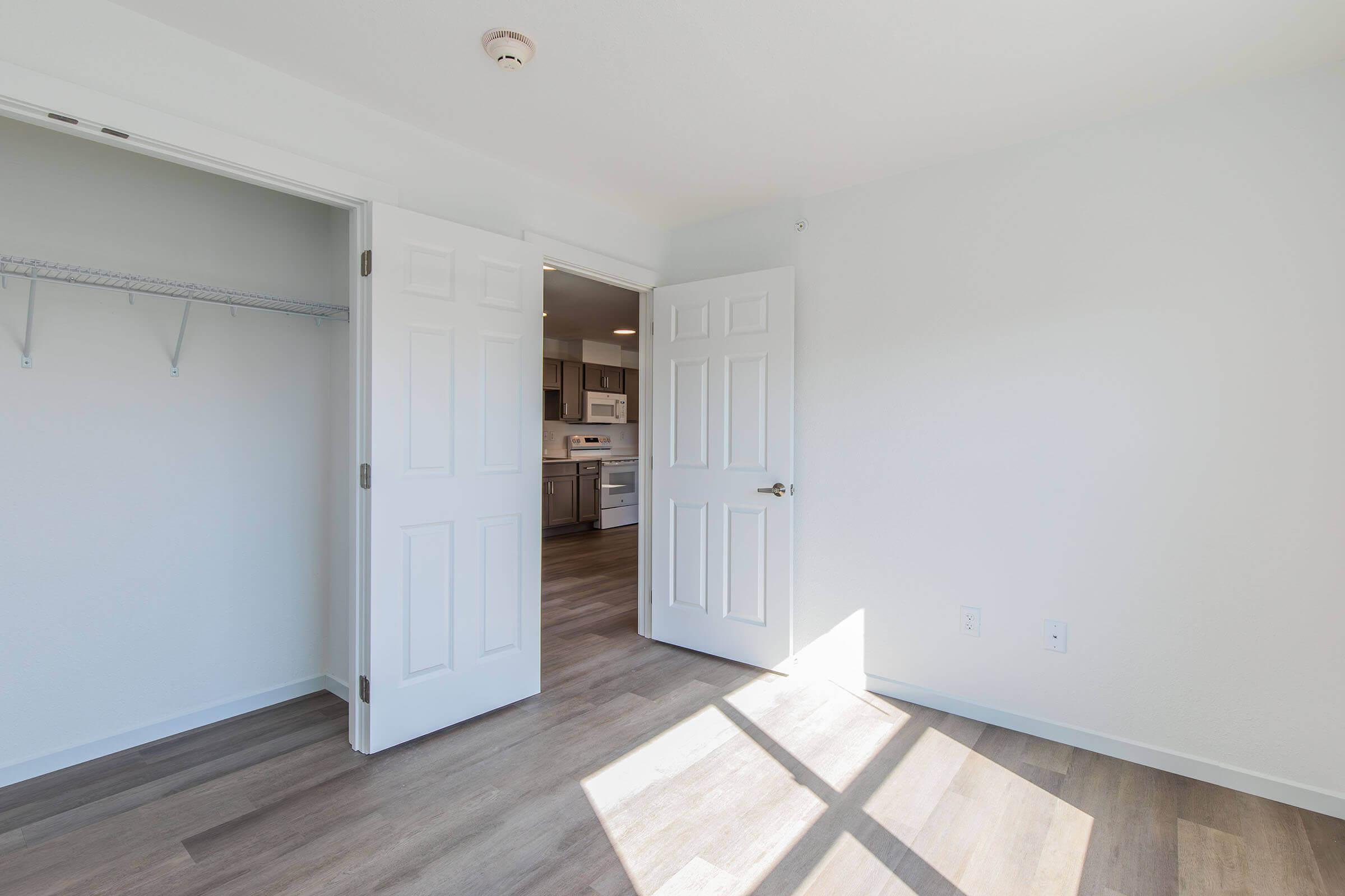 A bright, empty room with two open double doors leading to a kitchen area. The walls are painted white, and the floor is made of light-colored wood. A closet shelf is visible on the left side. Natural light streams in, creating a warm and inviting atmosphere.