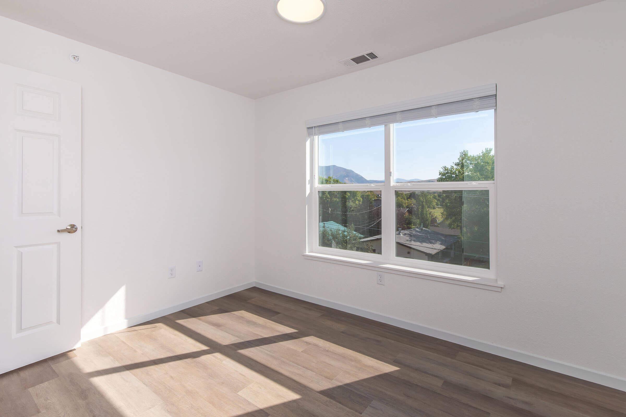 Bright, empty room with light-colored walls and a large window showing outdoor views. The floor is light wood, and sunlight casts shadows across the space. A door is visible on the left. Ideal for showcasing a minimalist living area or a potential bedroom.