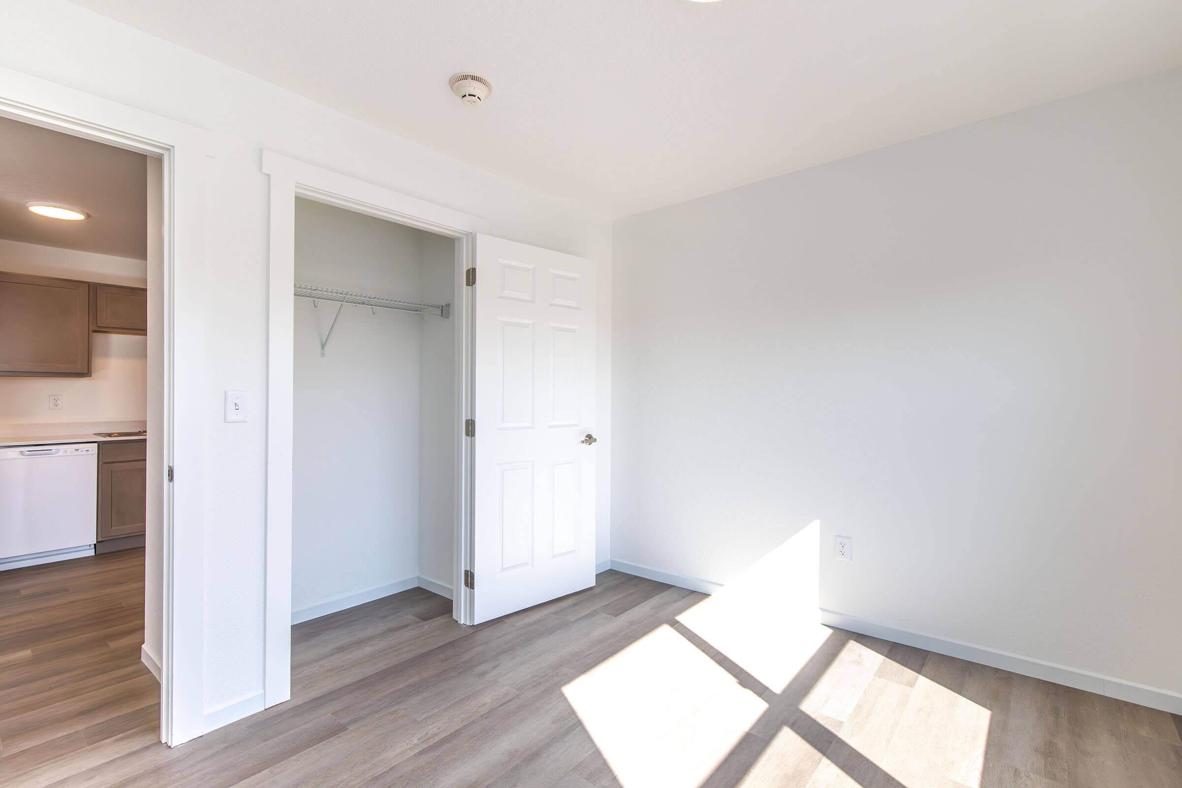 Interior view of a bright, modern room with light-colored walls and wood-style flooring. A white door leads to a closet, and another door opens to a space with kitchen appliances visible. Natural light streams in through a window, creating a welcoming atmosphere.