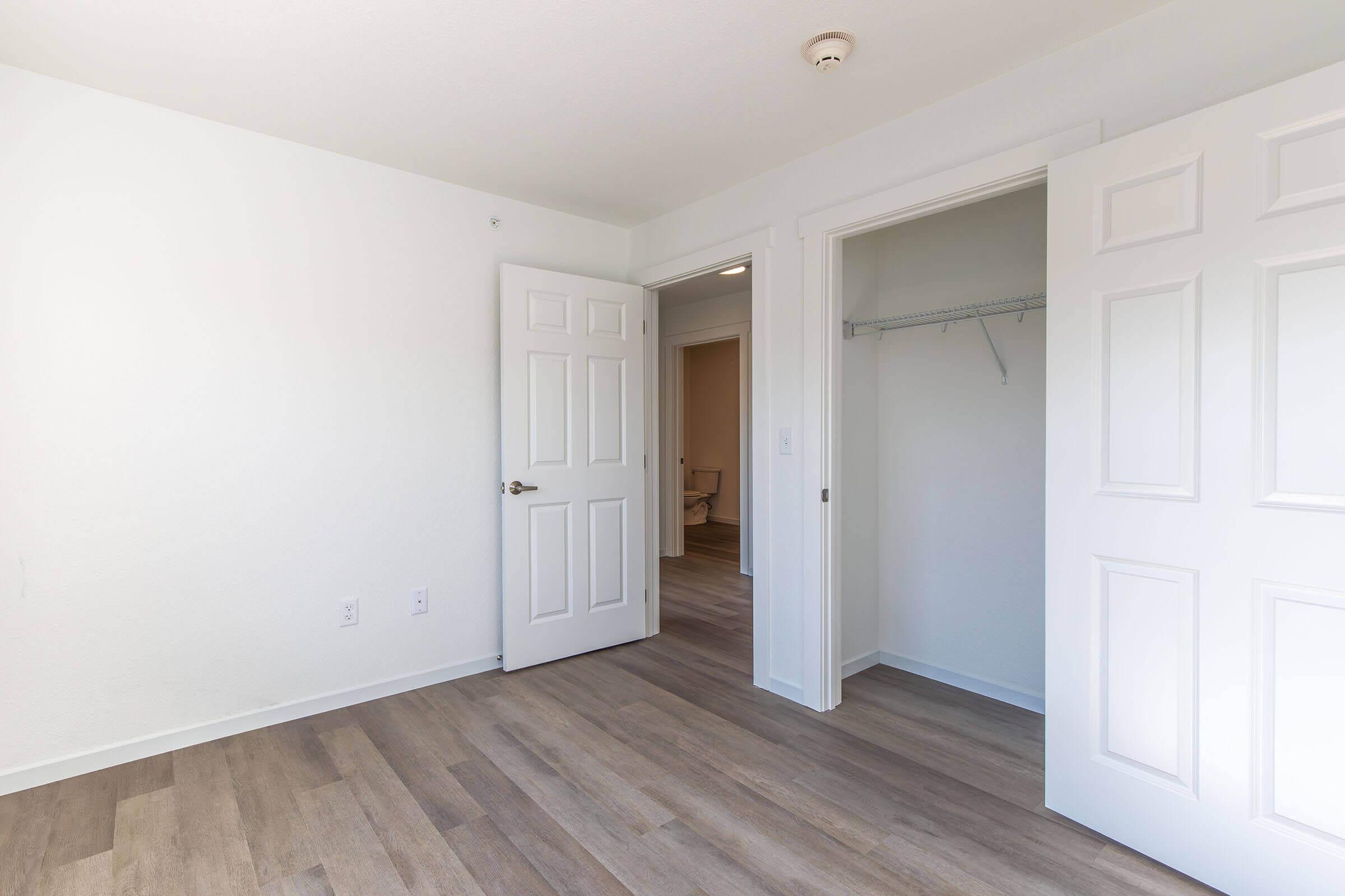 A bright, empty room with light-colored walls and wooden flooring. Two doors are visible: one open leading to another room and another to a closet. The space has a clean and modern aesthetic, with abundant natural light enhancing the openness of the area.
