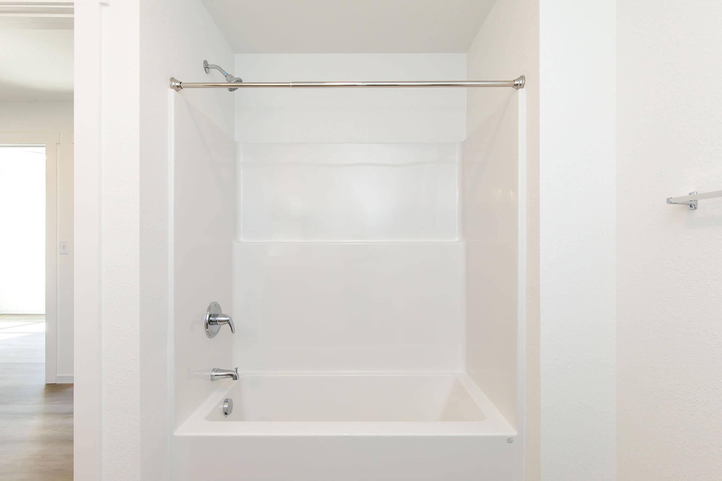 A clean, modern bathroom featuring a white bathtub with a smooth, glossy finish. The bathtub is enclosed by a clear shower curtain rod and a minimalistic design. The walls are painted white, providing a bright and spacious feel. Natural light can be seen entering the room from an adjacent door.