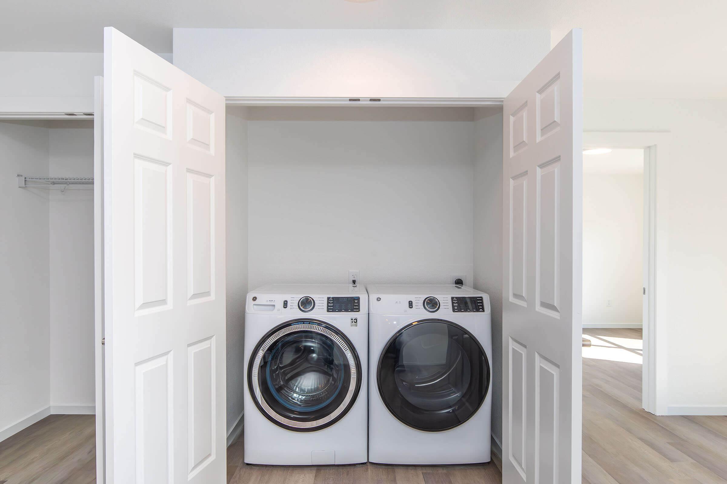 A modern laundry area featuring a stacked washer and dryer in a spacious closet with double doors. The room is well-lit with light-colored walls and wooden flooring, creating a clean and organized appearance.