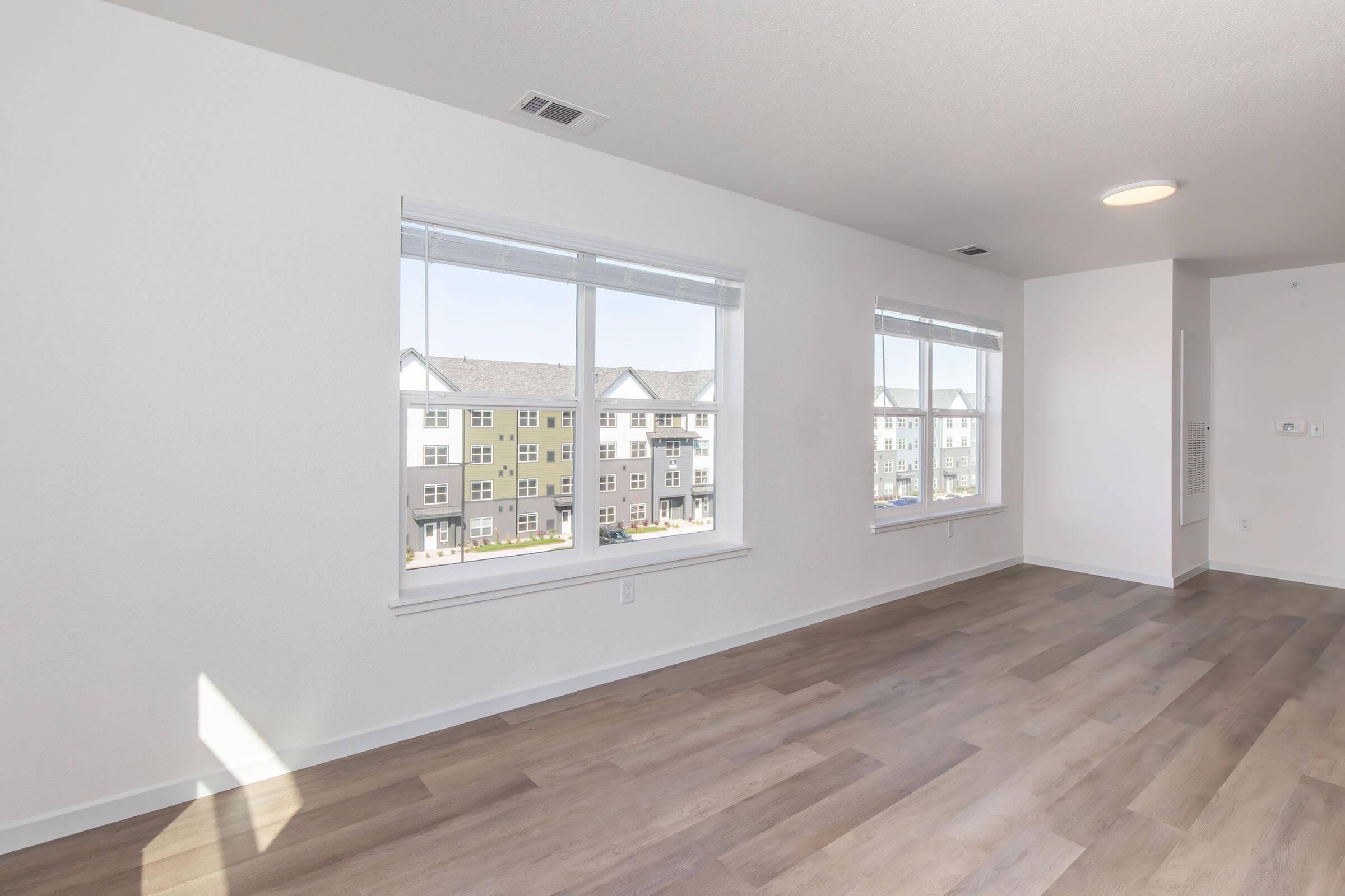 Spacious, bright interior of a modern room with large windows showcasing views of adjacent buildings. The floor features light-colored wood, and the walls are painted in a clean, neutral tone, enhancing the open and airy atmosphere.