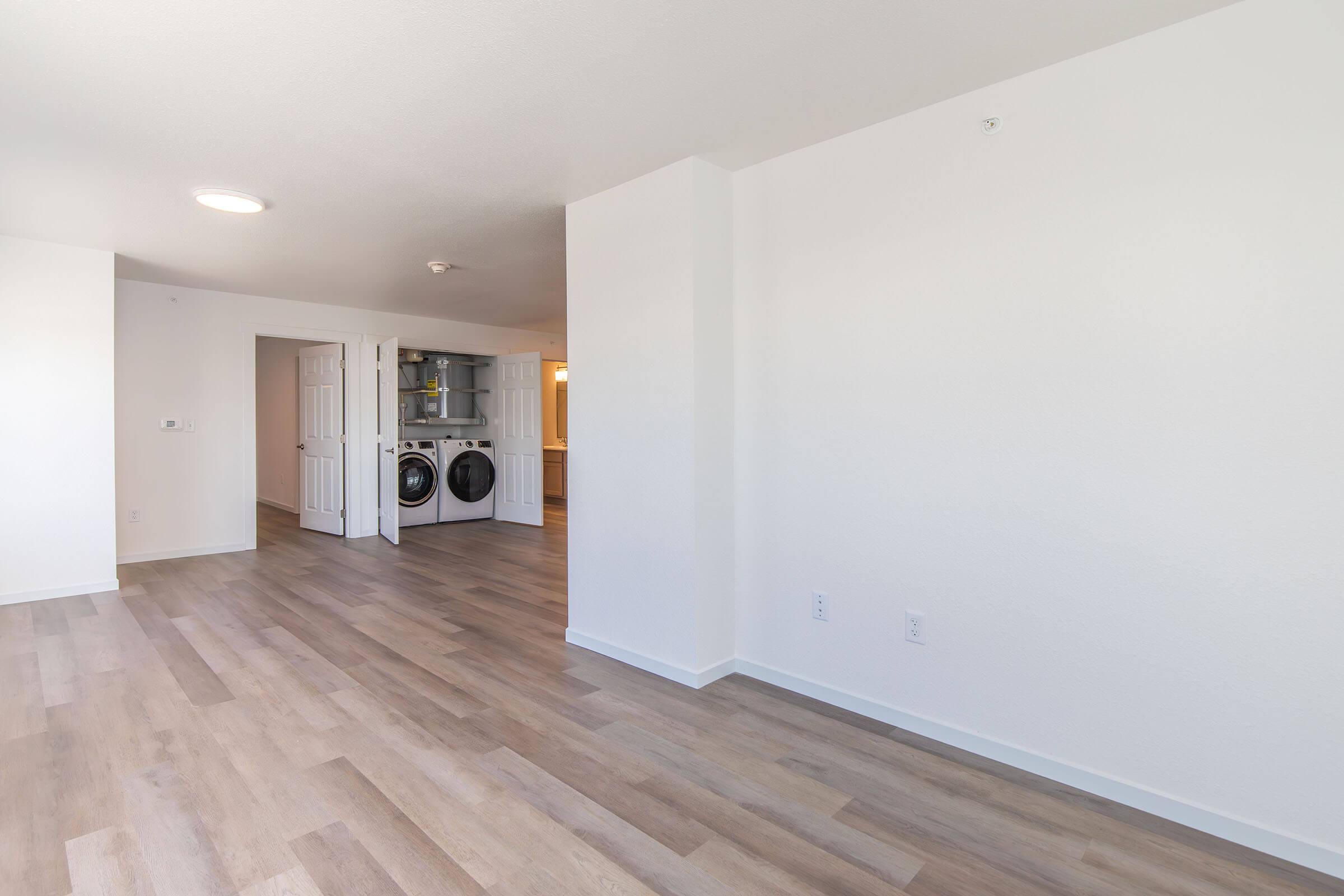 Bright interior of a modern apartment featuring light-colored walls and wooden flooring. The image shows a spacious living area adjacent to a laundry area with stacked appliances. Natural light fills the room, creating an inviting atmosphere.