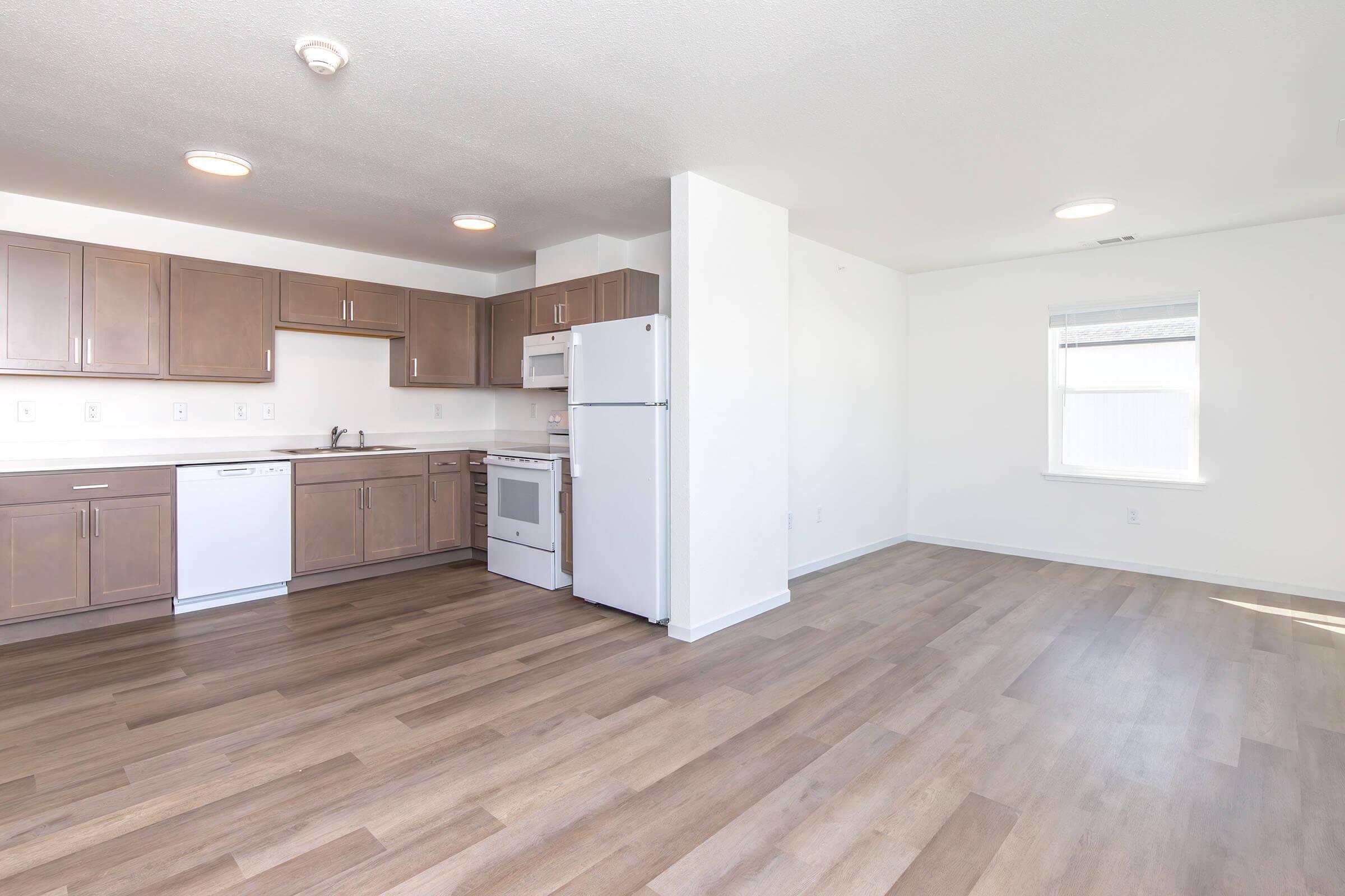 Modern kitchen with wooden cabinets, white appliances including a dishwasher and oven, and a spacious living area with light-colored flooring. Natural light enters through a window, creating a bright and inviting atmosphere.