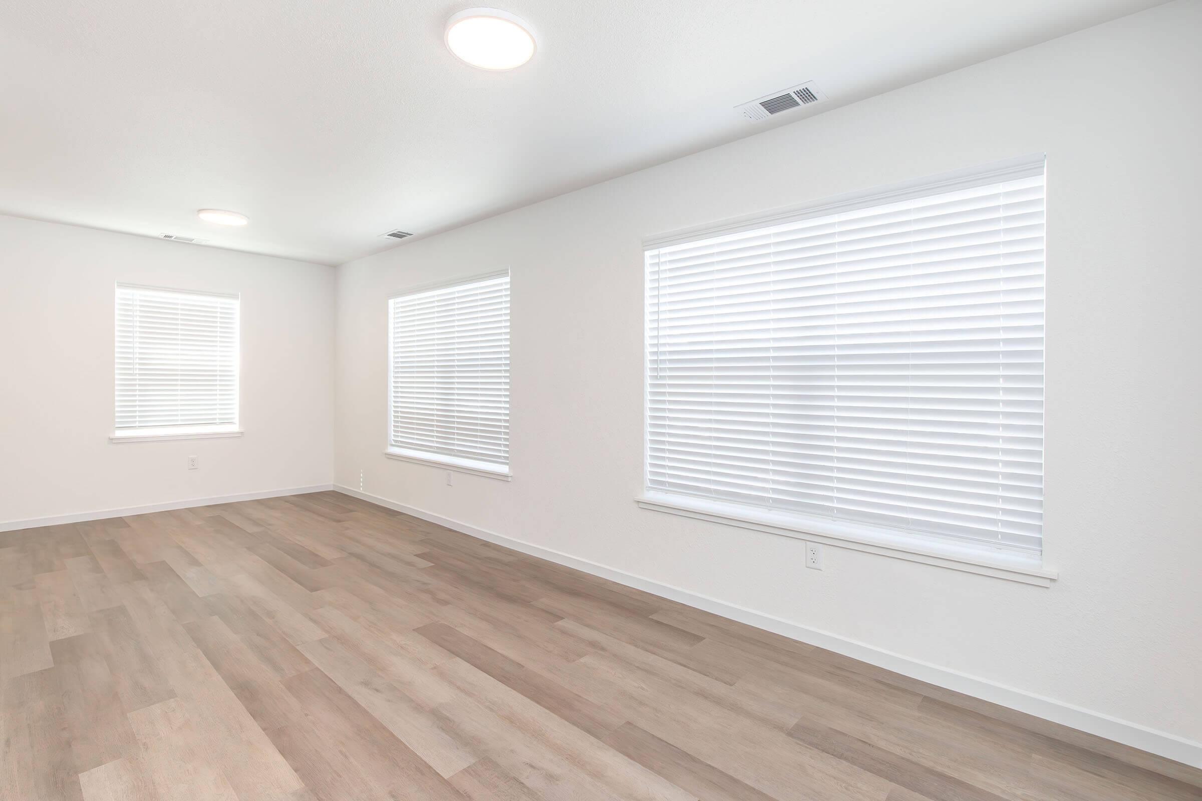 A bright, empty room with white walls and two large windows covered with white blinds. The flooring is light-colored wood, and there are recessed ceiling lights providing ample illumination. The space feels open and airy, ideal for various uses or decoration.