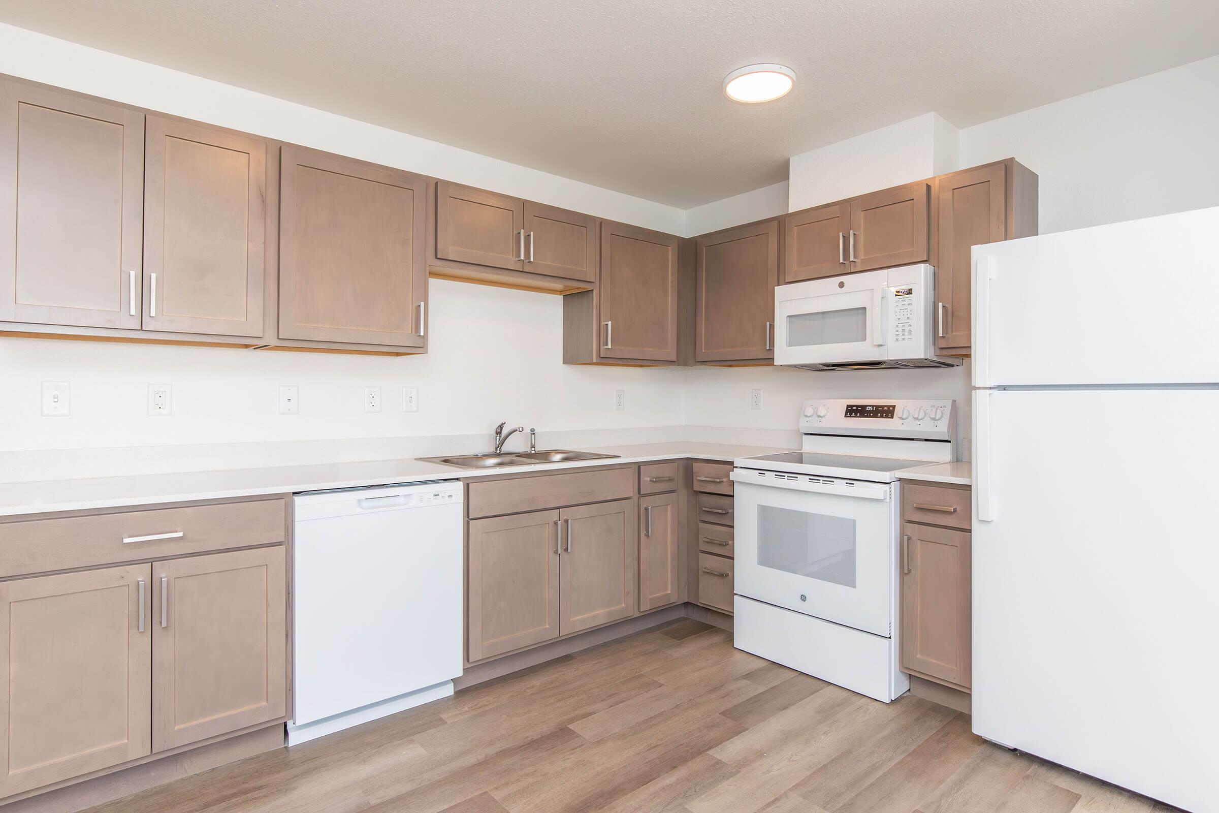 Modern kitchen featuring light brown cabinets, white countertops, and stainless steel appliances. Includes a double sink, dishwasher, built-in microwave, and oven. The space is bright with natural light and has a clean, contemporary design.