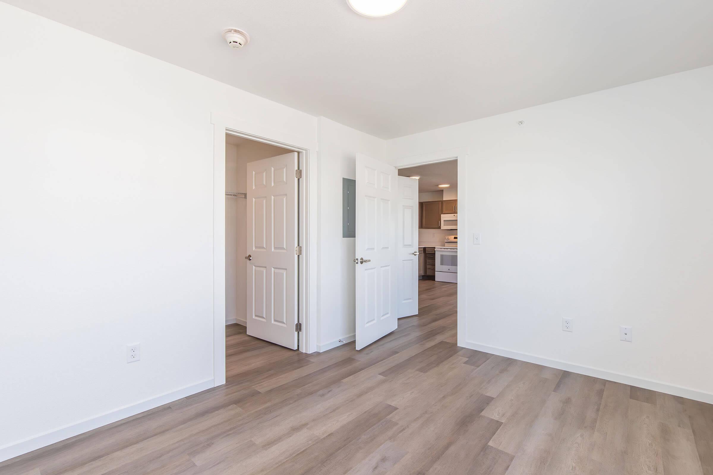 A spacious room with light-colored walls and wooden flooring. Two doors are visible on the left, leading to a closet and another room. In the background, a kitchen area is partially visible, featuring modern appliances and cabinetry. The overall ambiance is bright and contemporary.