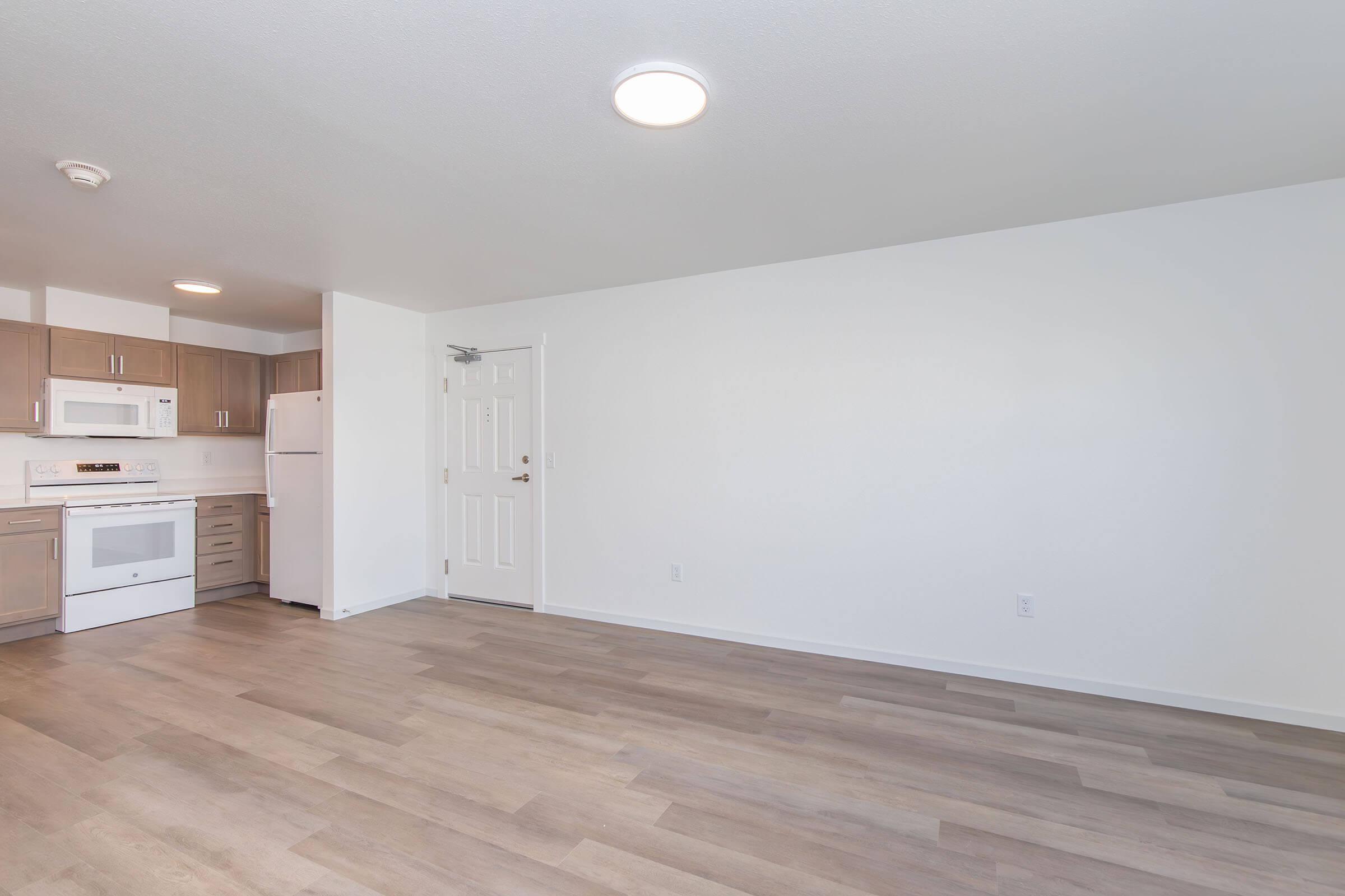 A modern, spacious kitchen area with light wood flooring, white walls, and a ceiling light. The kitchen features beige cabinetry, a microwave, a refrigerator, and an oven, with no clutter in the open living space, creating a bright and airy atmosphere.