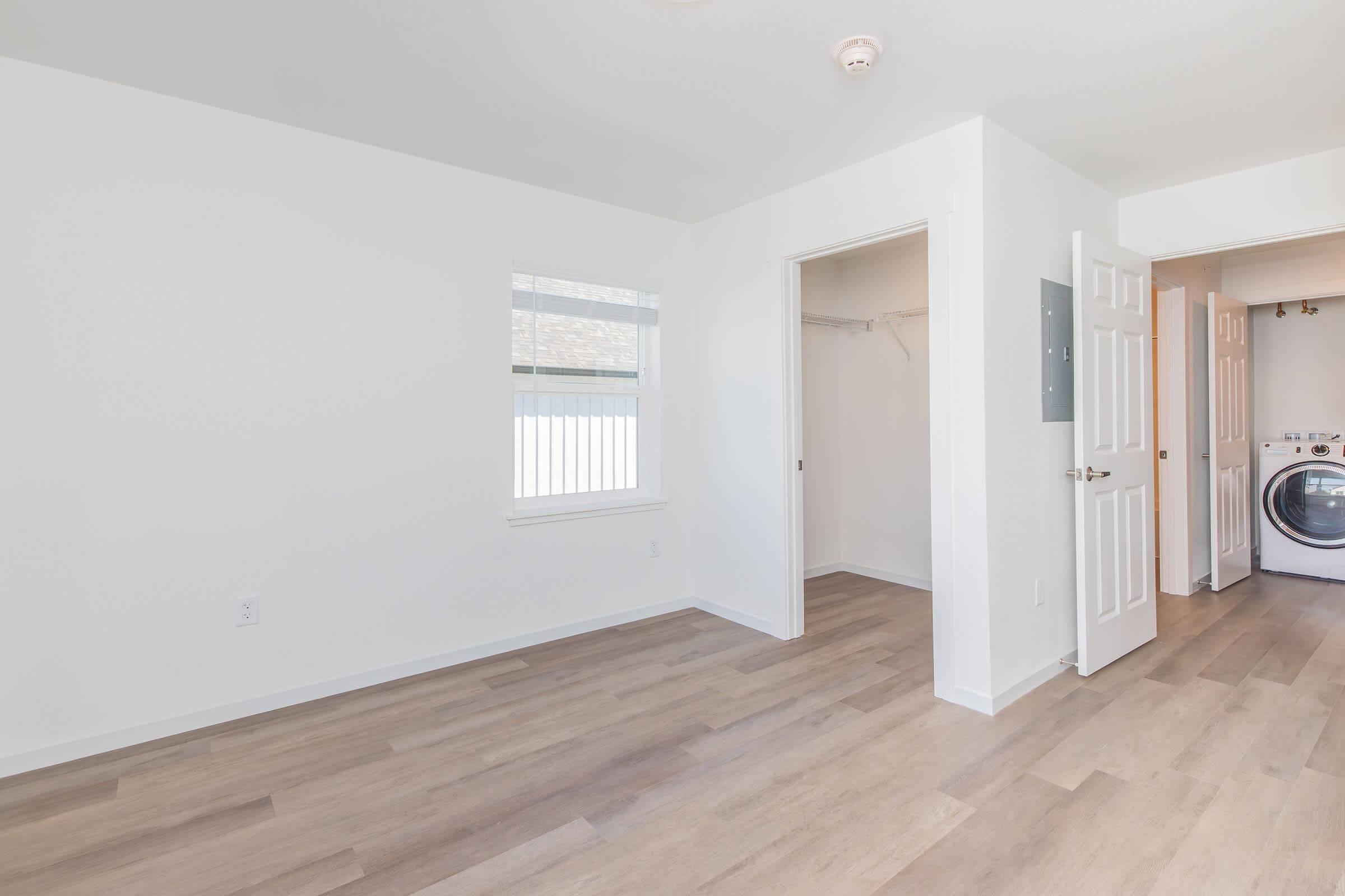 A bright, empty room with light-colored walls and wood-like flooring. One side features a window, while the opposite side has an open closet space and a laundry area with a washer and dryer. The overall aesthetic is clean and modern, creating a spacious feel.