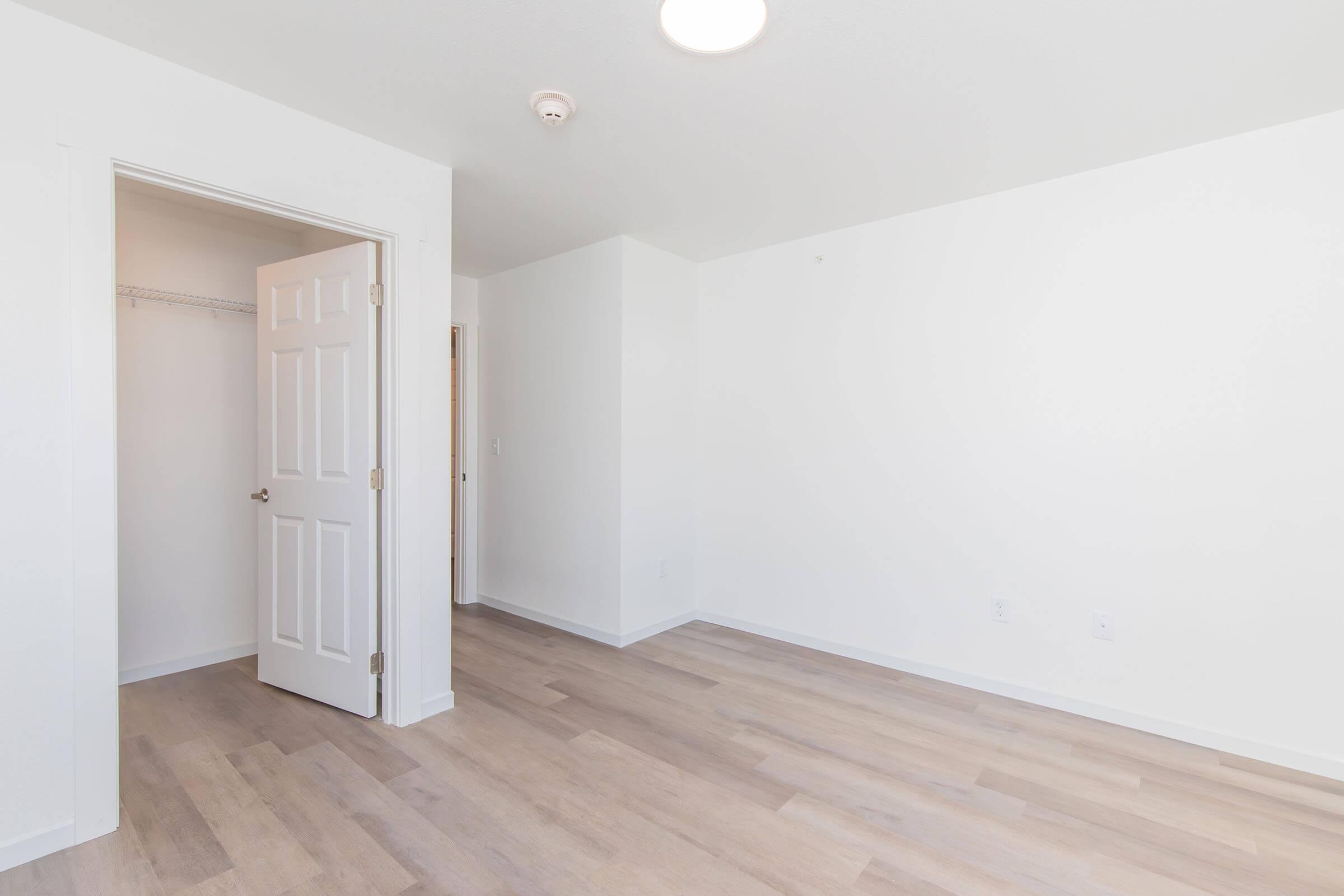 Empty room with light-colored walls and a wooden floor, featuring an open door leading to a closet. The space is bright and unfurnished, with a ceiling light fixture visible. There are no decorations or furniture in the room, emphasizing a minimalist aesthetic.