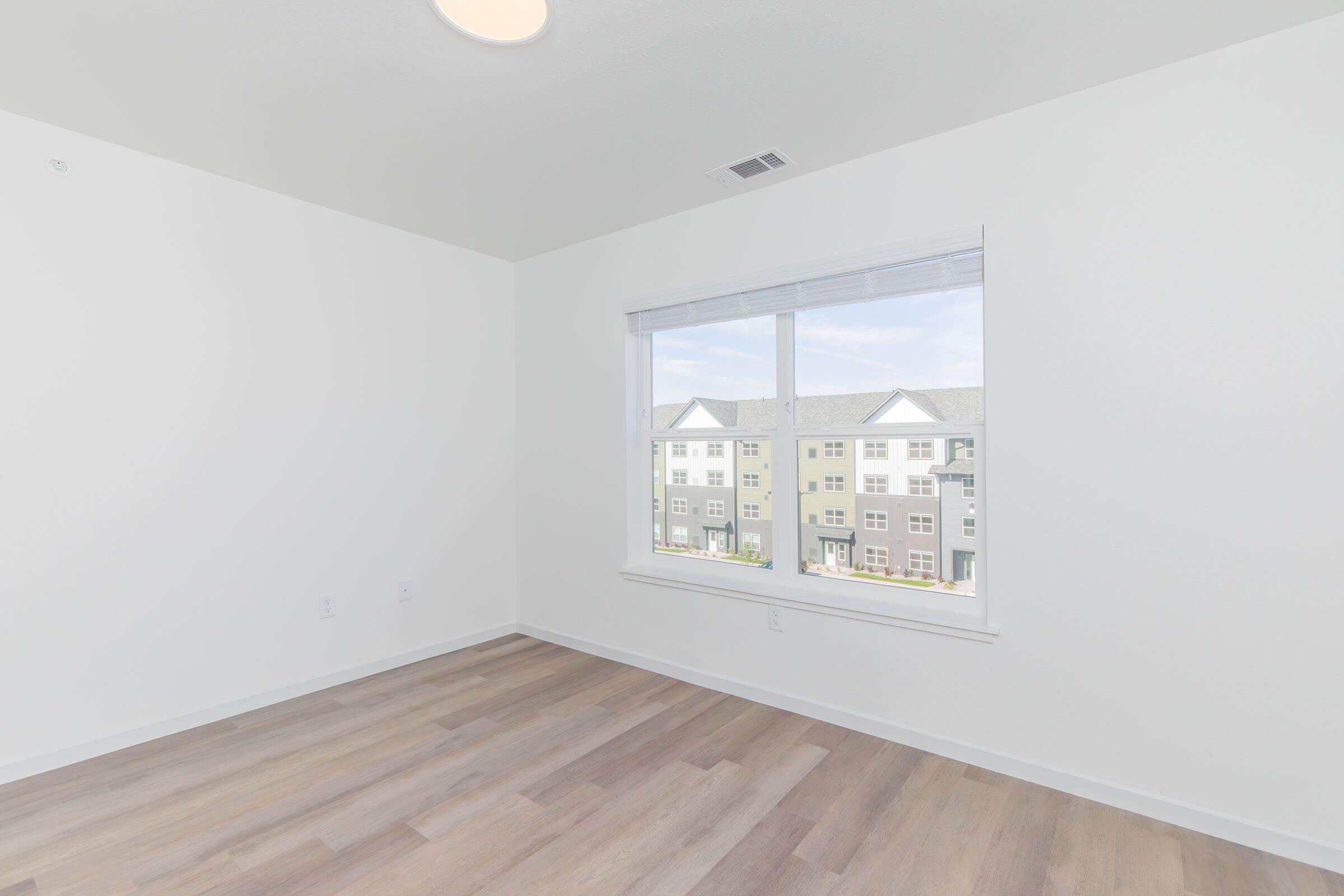 Empty room with light-colored walls and a large window facing an exterior view. Natural light floods the space, highlighting the wooden floor. The window offers a view of an apartment complex across the street. The ceiling features a circular light fixture.