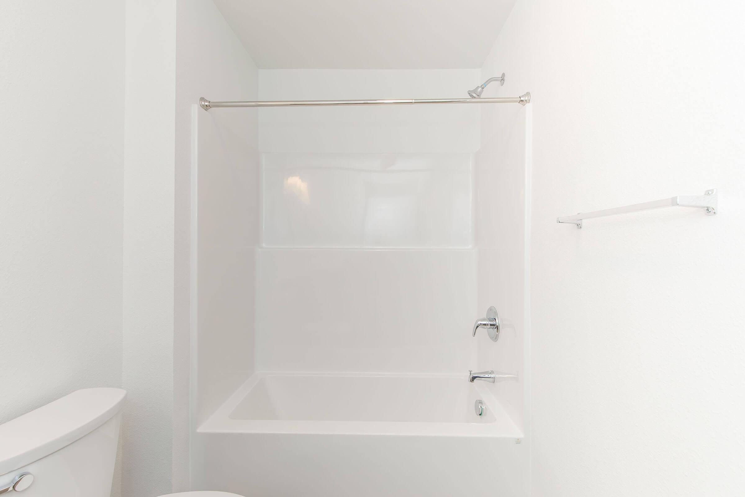 A clean, modern bathroom showing a white bathtub with a glass shower curtain rod. The wall and surrounding area are painted white, and there is a chrome faucet and showerhead. On the left side, a white toilet is partially visible, while a towel bar is mounted on the wall.