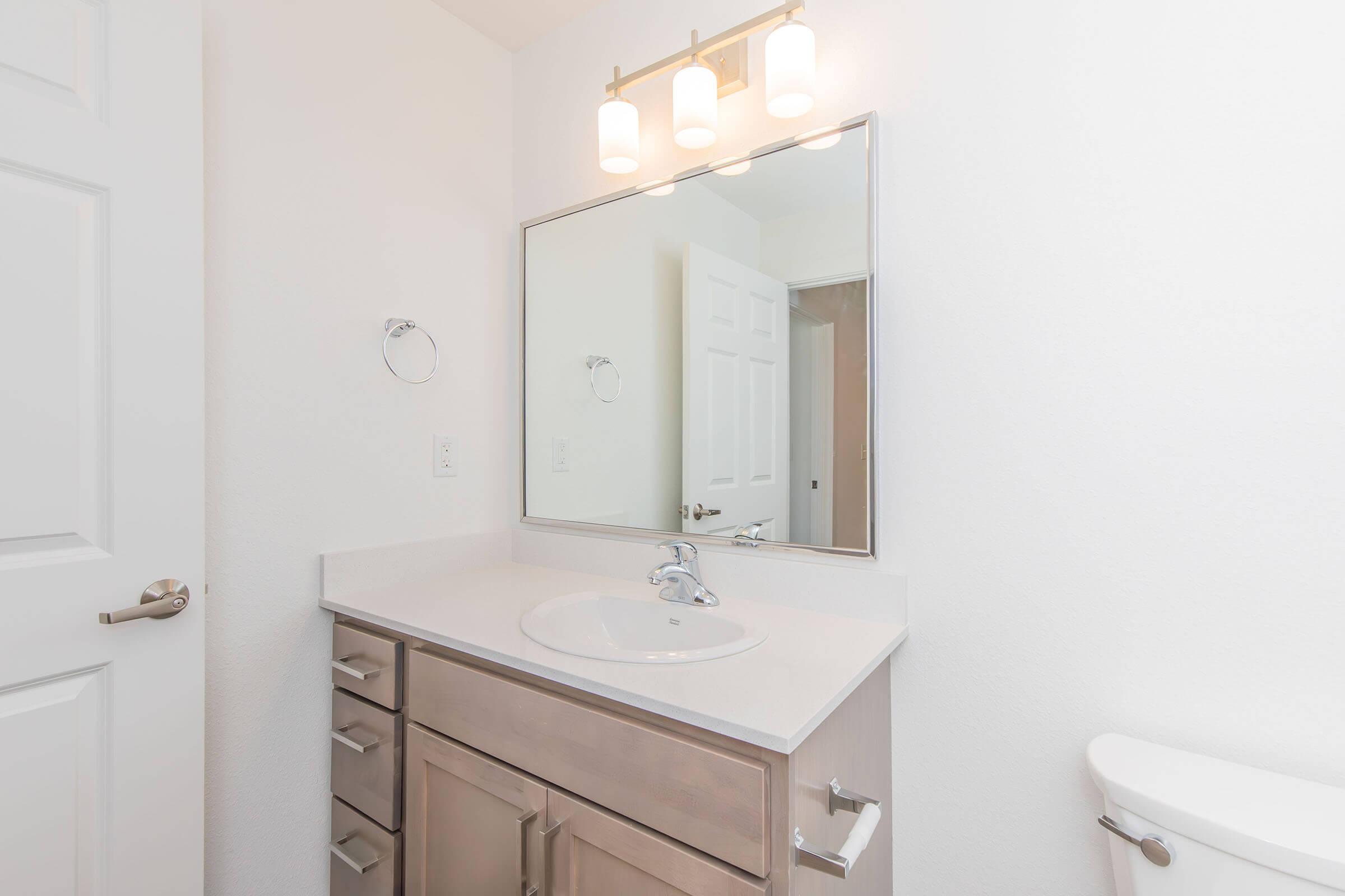 A clean, modern bathroom featuring a white sink with a chrome faucet, a large mirror above it, and three pendant lights. Light-colored cabinets and a towel holder are visible, along with a white toilet and a closed door in the background, giving the space a bright and airy feel.