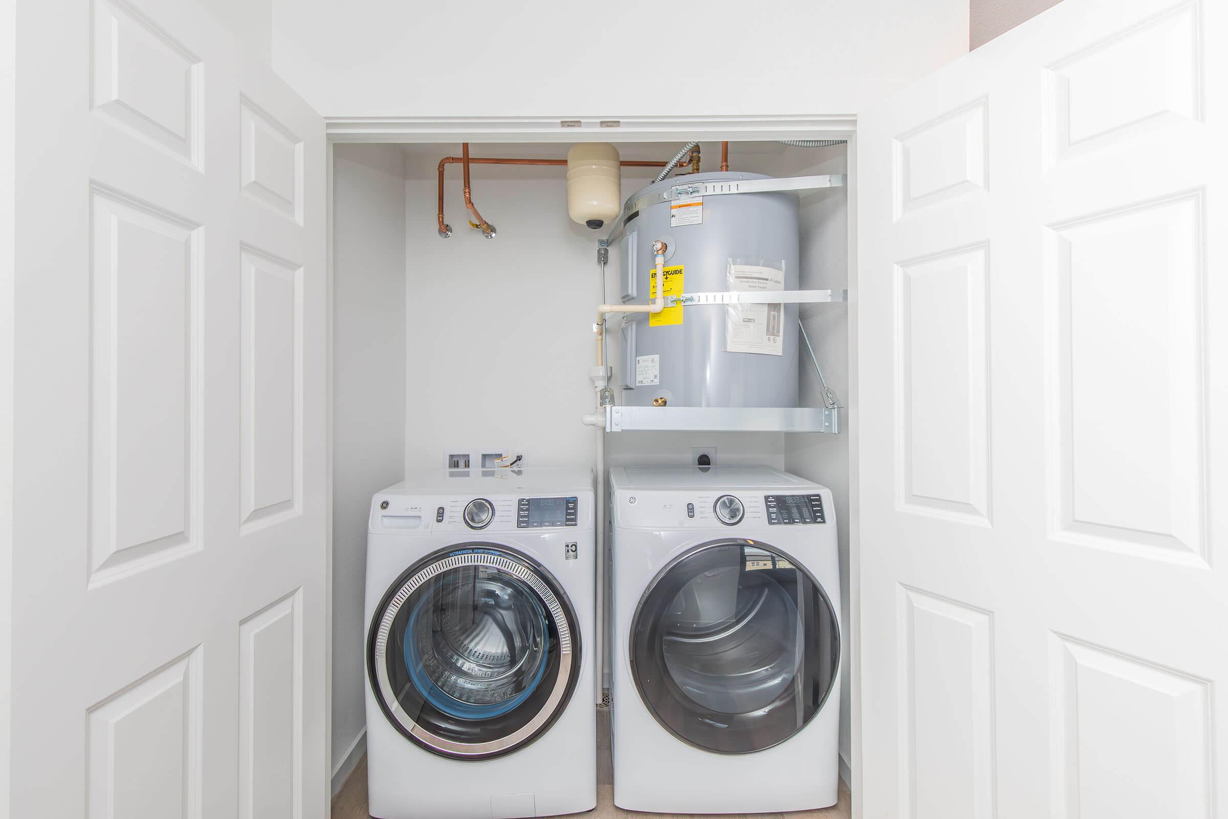 A laundry closet featuring a white stackable washer and dryer side by side, with a water heater mounted above. The doors are closed, and the walls are painted light colors, creating a clean and organized look. Various plumbing and utility connections are visible.