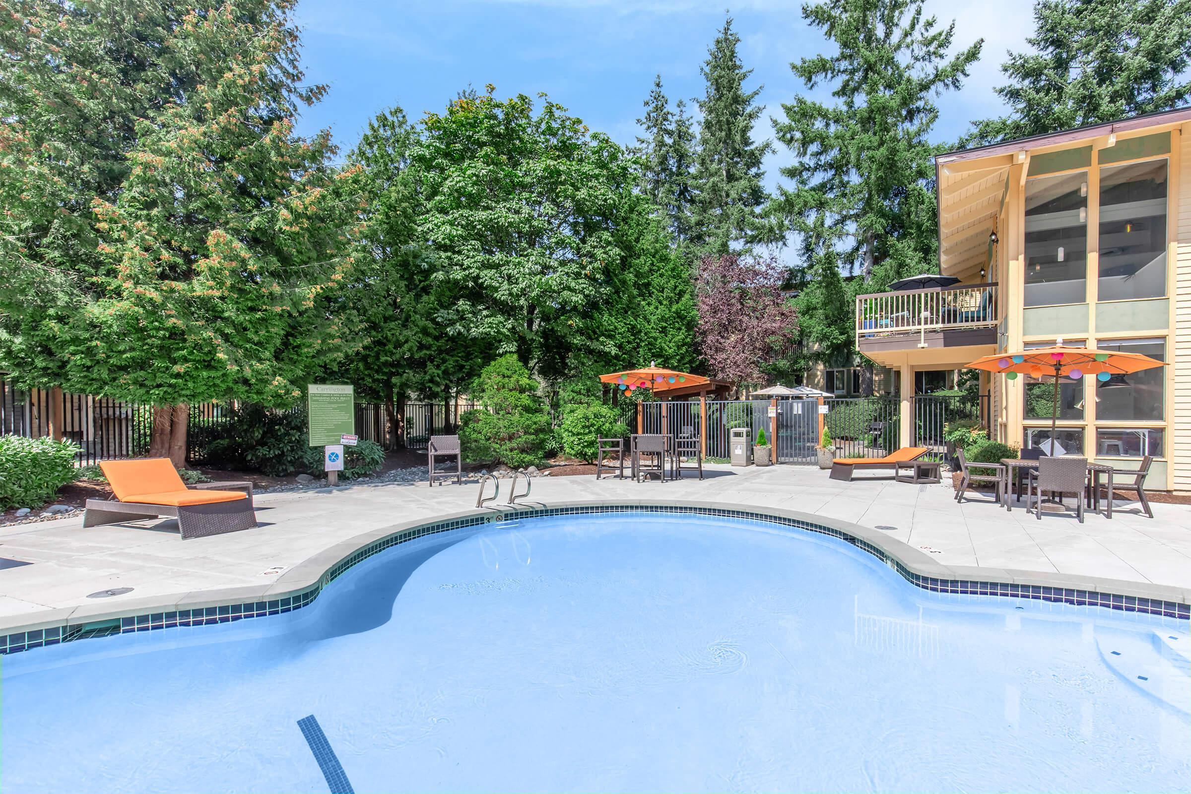 A serene outdoor pool area with clear blue water, surrounded by lounge chairs and shaded tables under orange umbrellas. Lush green trees and landscaping create a tranquil atmosphere, while a modern building with a balcony is visible in the background.
