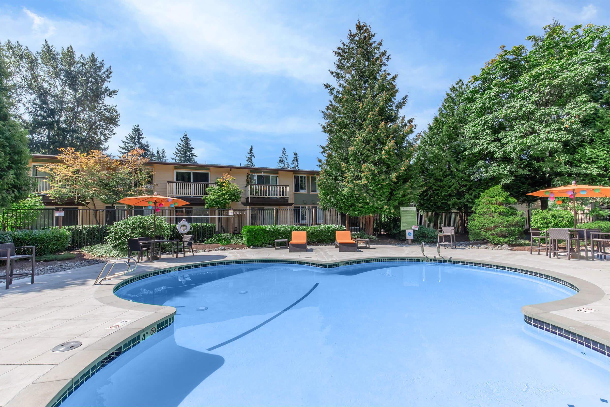 A serene view of a swimming pool surrounded by lush greenery, with orange umbrellas set up on wooden lounge chairs. In the background, there are well-maintained apartments with balconies, creating a relaxing outdoor atmosphere. Bright blue sky with a few clouds adds to the tranquil setting.