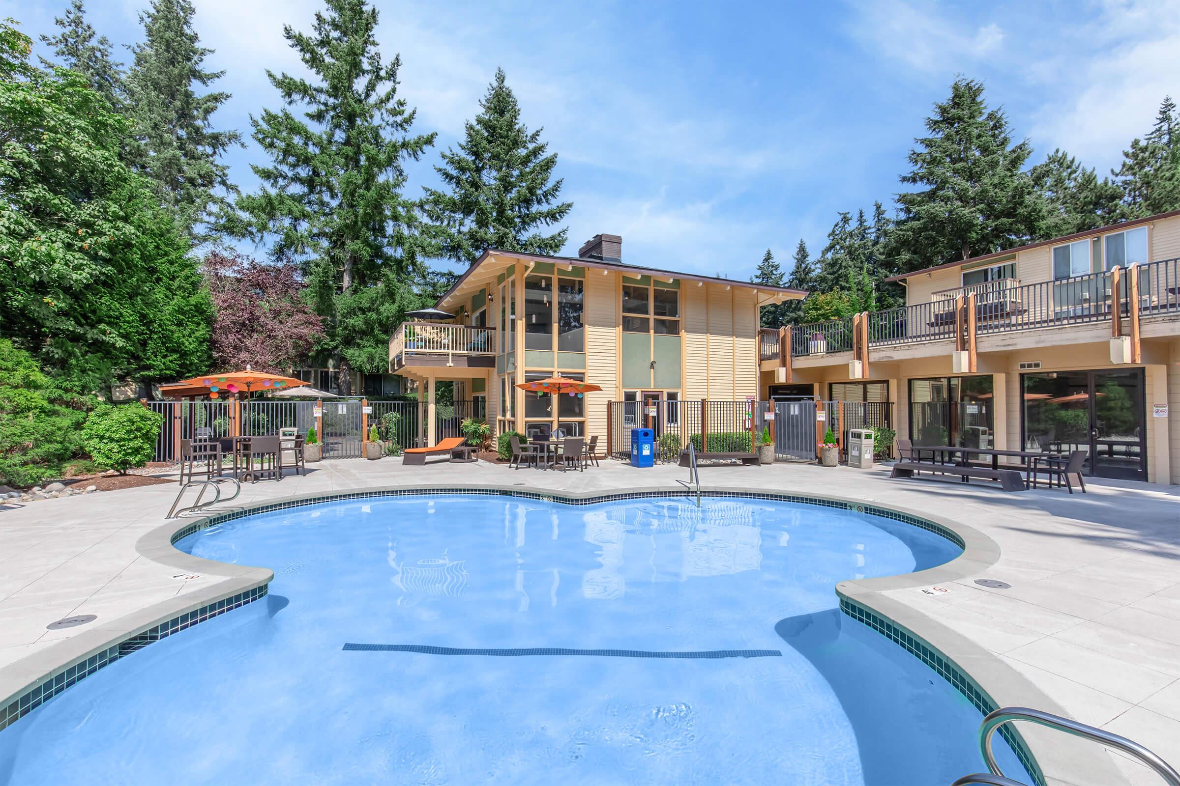 A swimming pool surrounded by a landscaped area, with lounge chairs and orange umbrellas. In the background, a multi-story building with balconies and surrounding greenery, including tall trees. The sky is clear and blue, creating a sunny atmosphere.