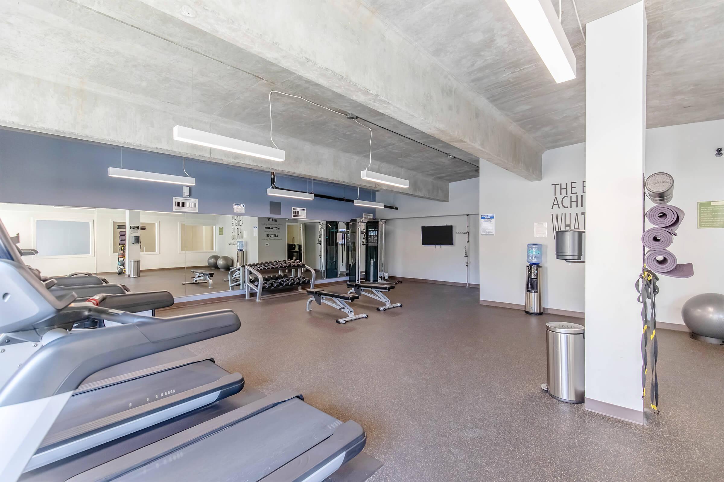 A spacious gym featuring treadmills, weightlifting equipment, and exercise mats. Large mirrors reflect the interior, and a water cooler is visible in the corner. The walls are painted in neutral colors with motivational quotes and fitness information displayed. Clean and well-lit environment.