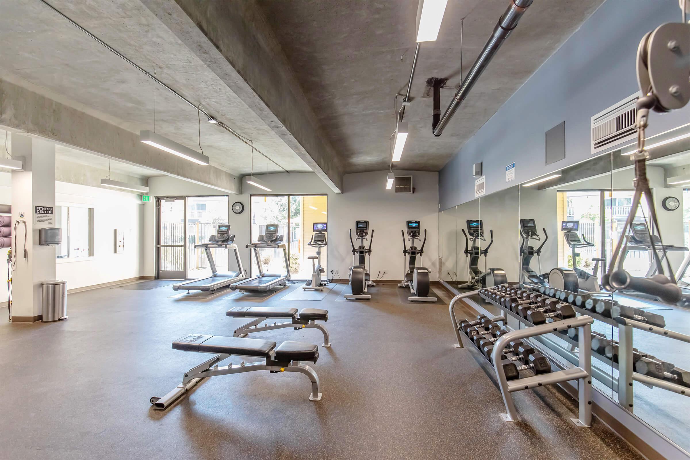A modern gym interior featuring several cardio machines including treadmills and elliptical trainers, along with weight benches and free weights. The space has a concrete ceiling, large mirrors, and ample natural light coming from windows.