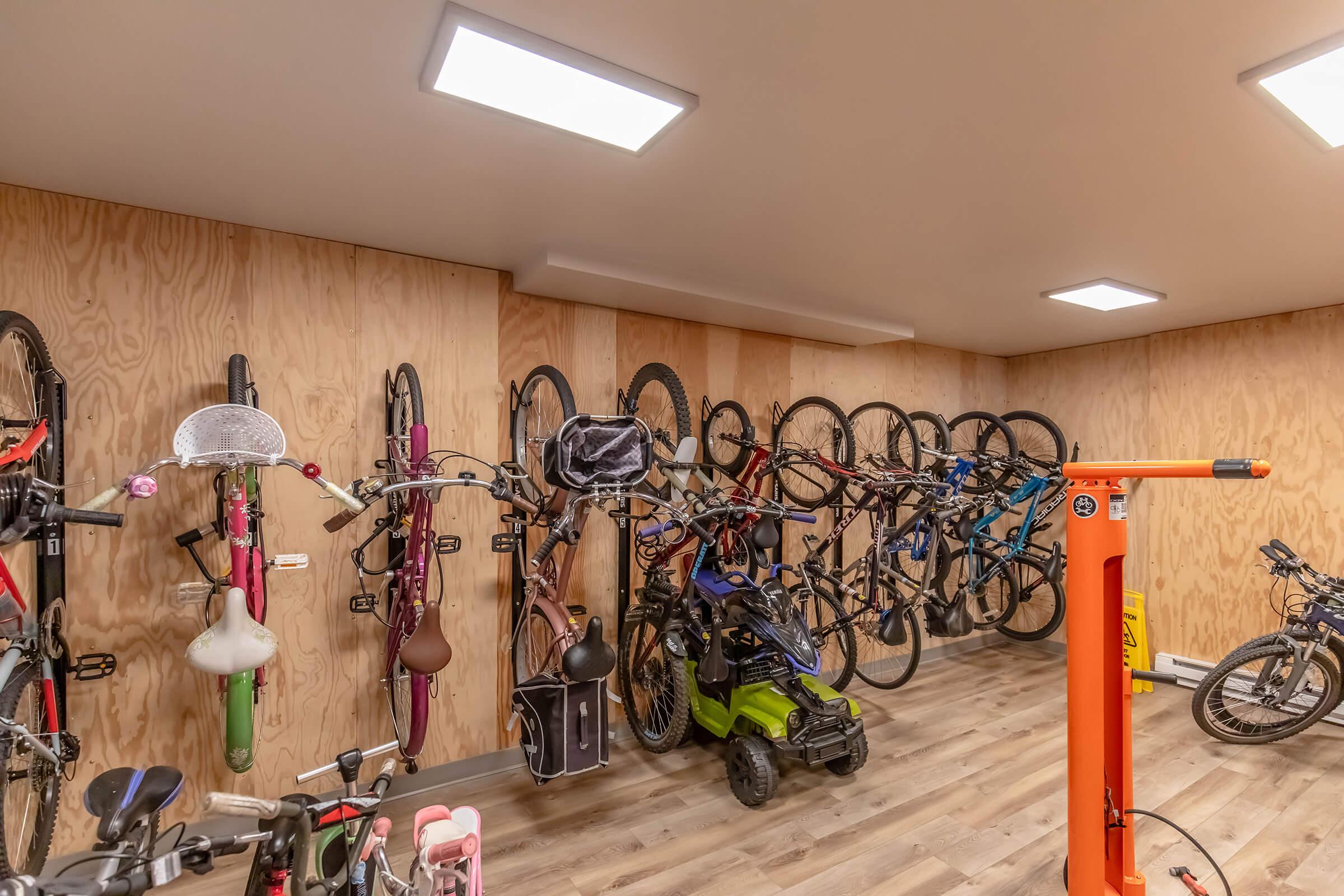 A well-organized indoor bike storage area featuring multiple bicycles mounted on wooden walls. The space has bright overhead lights, wooden flooring, and a bike repair station with tools in view. Various bike types and colors are visible, alongside a bike trailer.