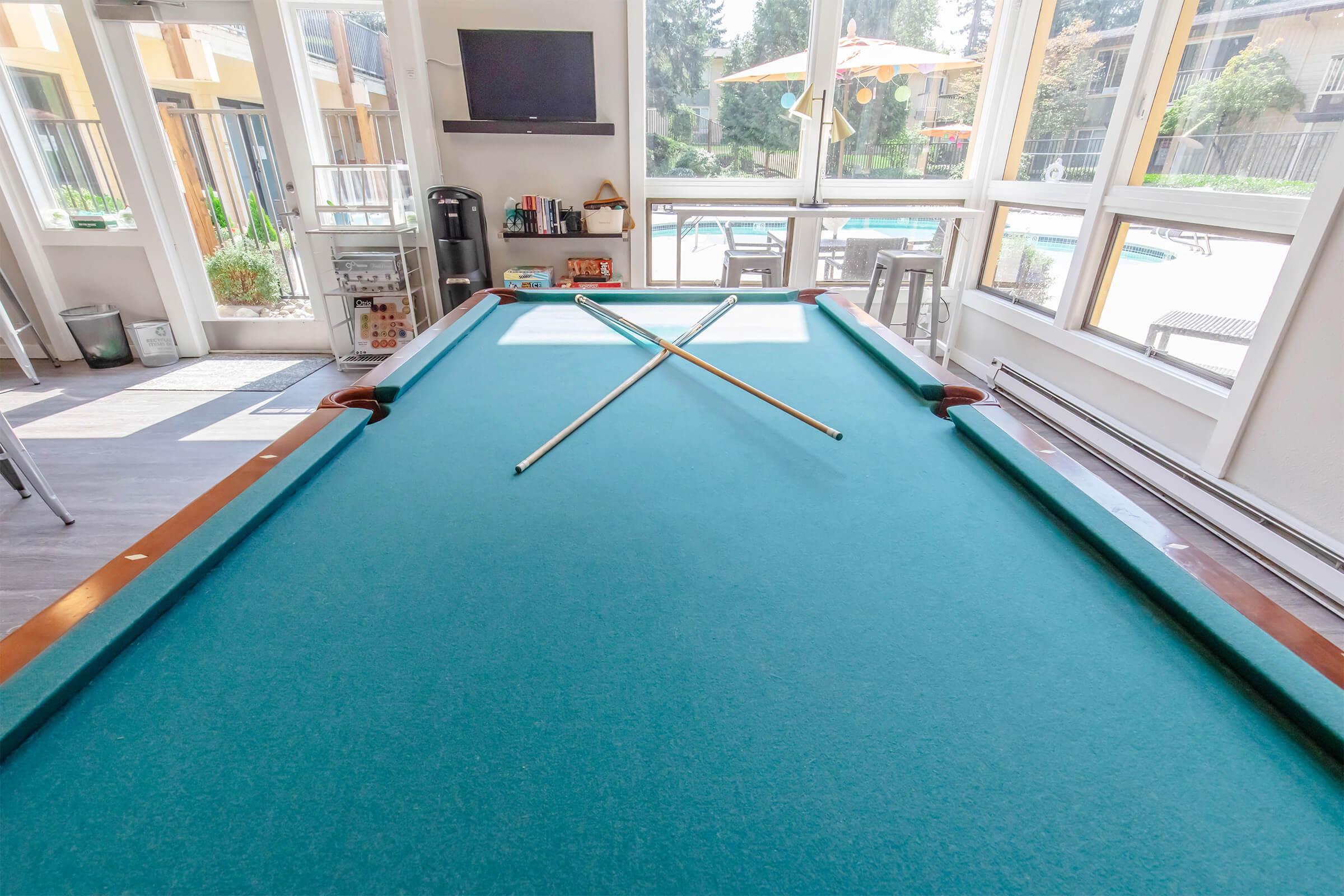 A well-lit room featuring a green felt pool table with two cue sticks crossed on top. Large windows allow natural light and provide a view of a pool area outside. A television is mounted on the wall, and the space is decorated with modern furniture and plants.